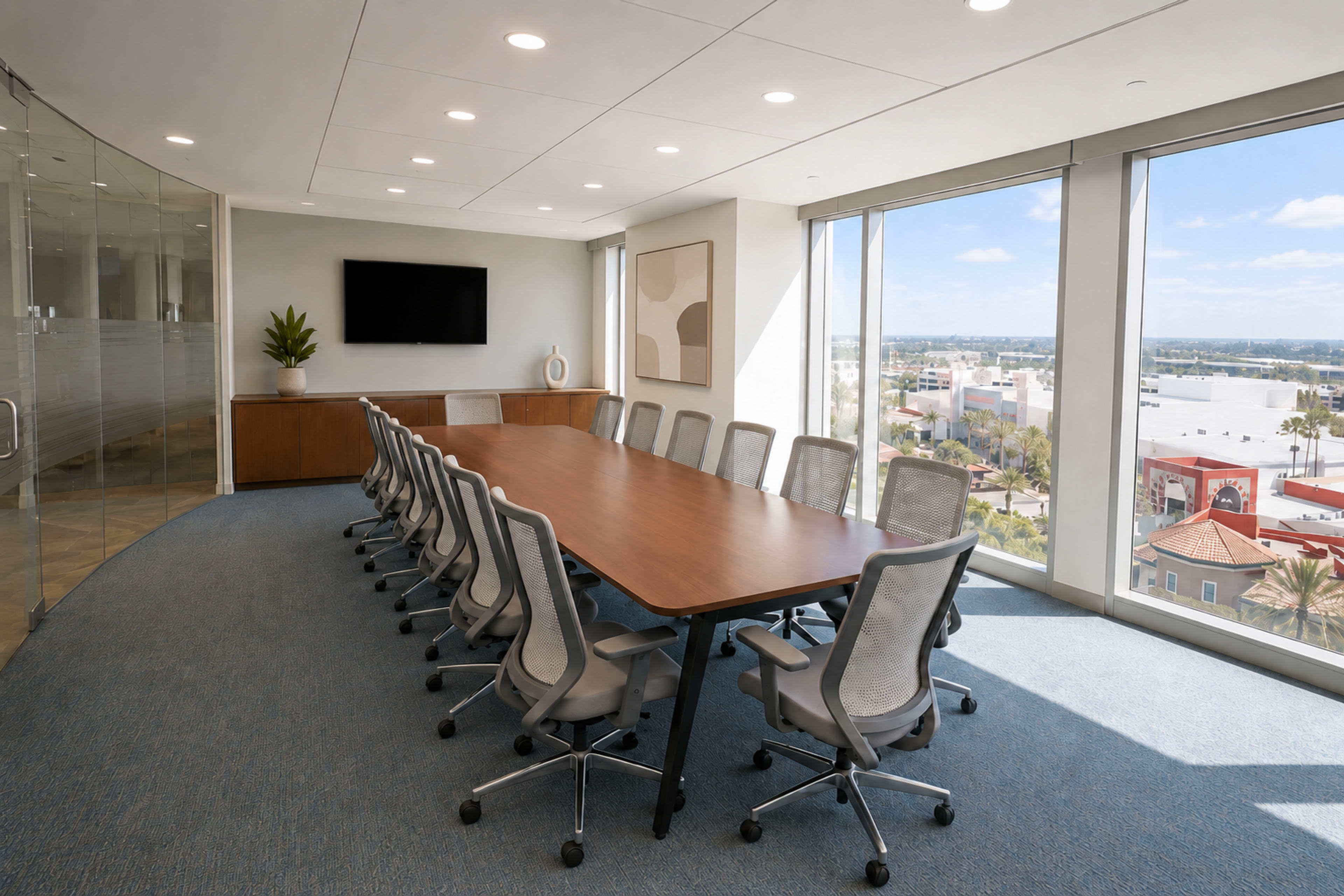 A spacious conference room features a large rectangular table surrounded by ergonomic chairs, with a wall-mounted screen and windows offering an outside view.