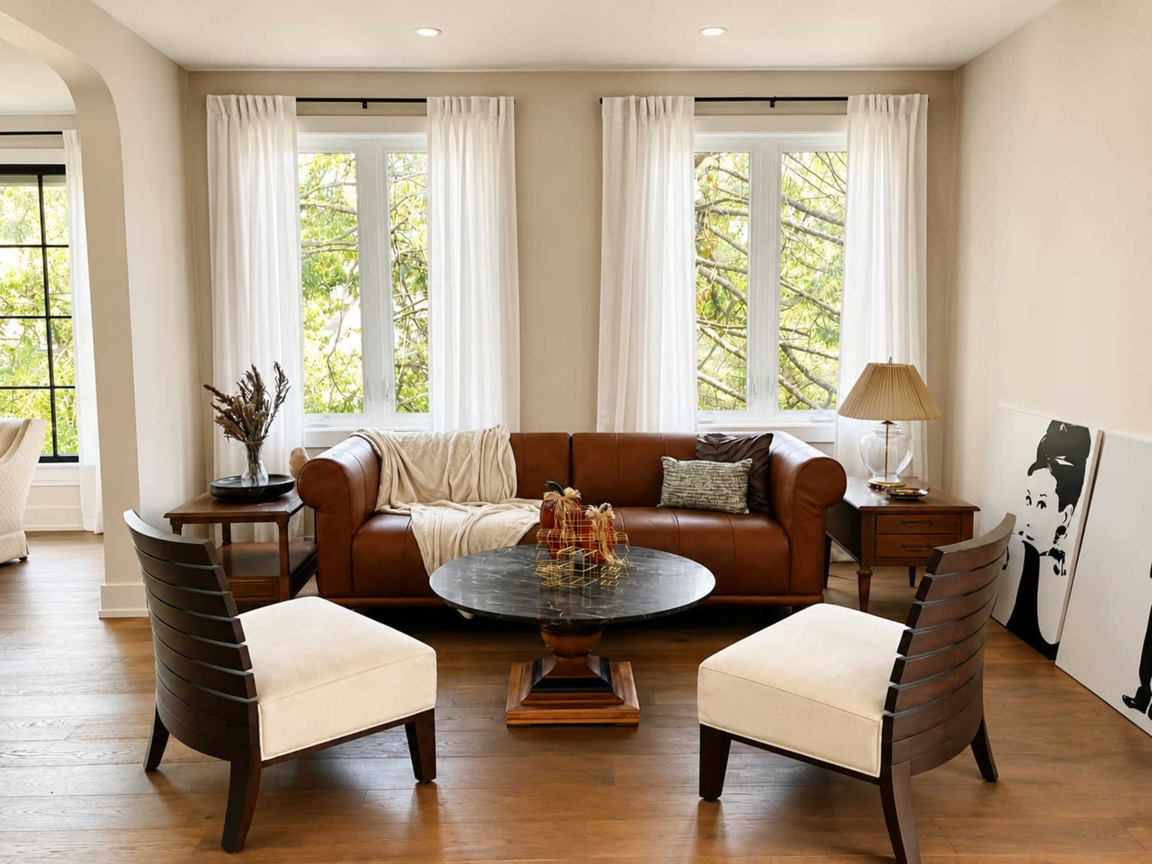 A cozy living room features a brown leather sofa, two upholstered chairs, a round coffee table, and large windows with white curtains letting in natural light.