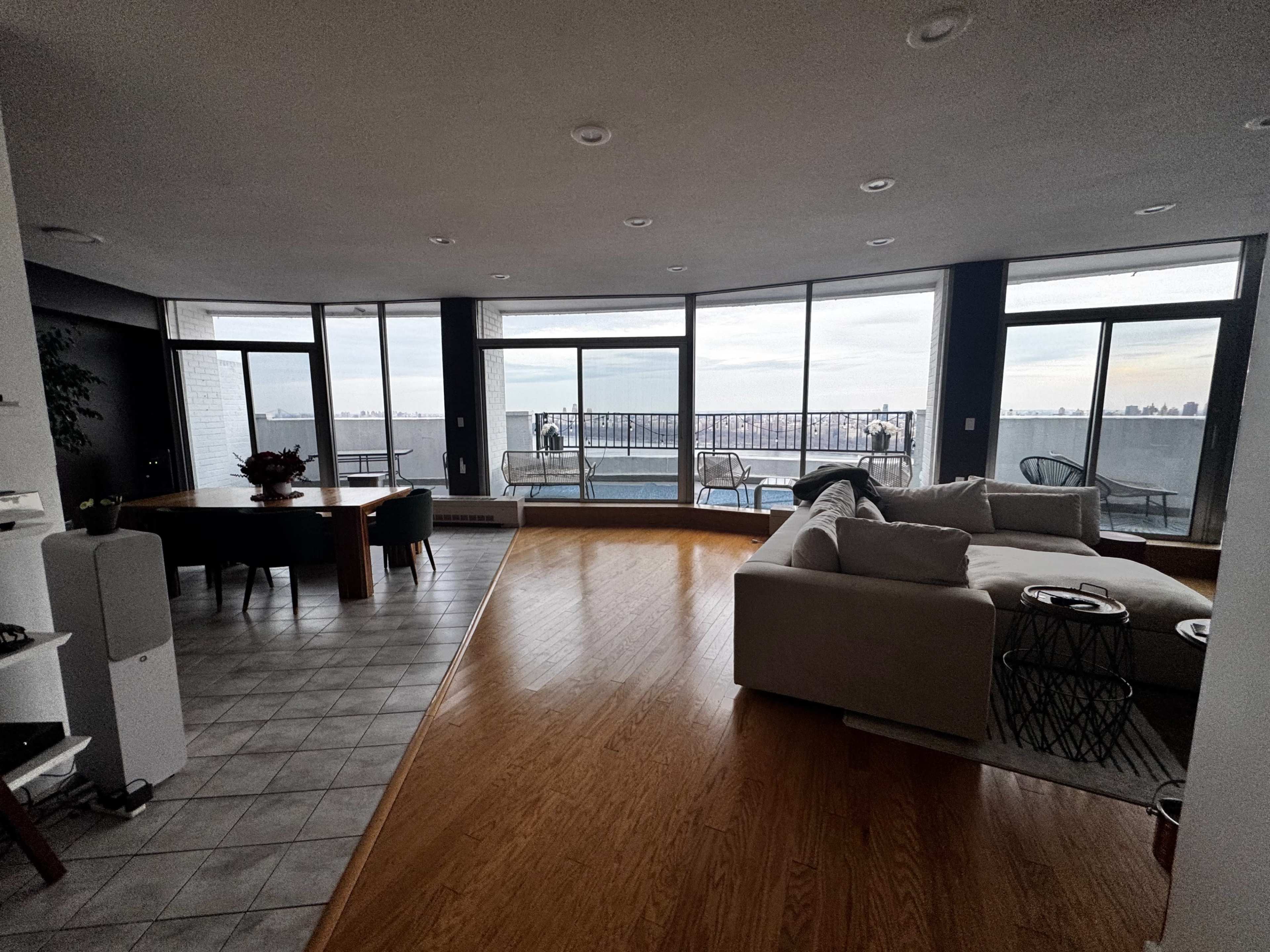 The image shows a spacious living area with large windows overlooking a body of water, featuring a dining table and a sectional sofa.