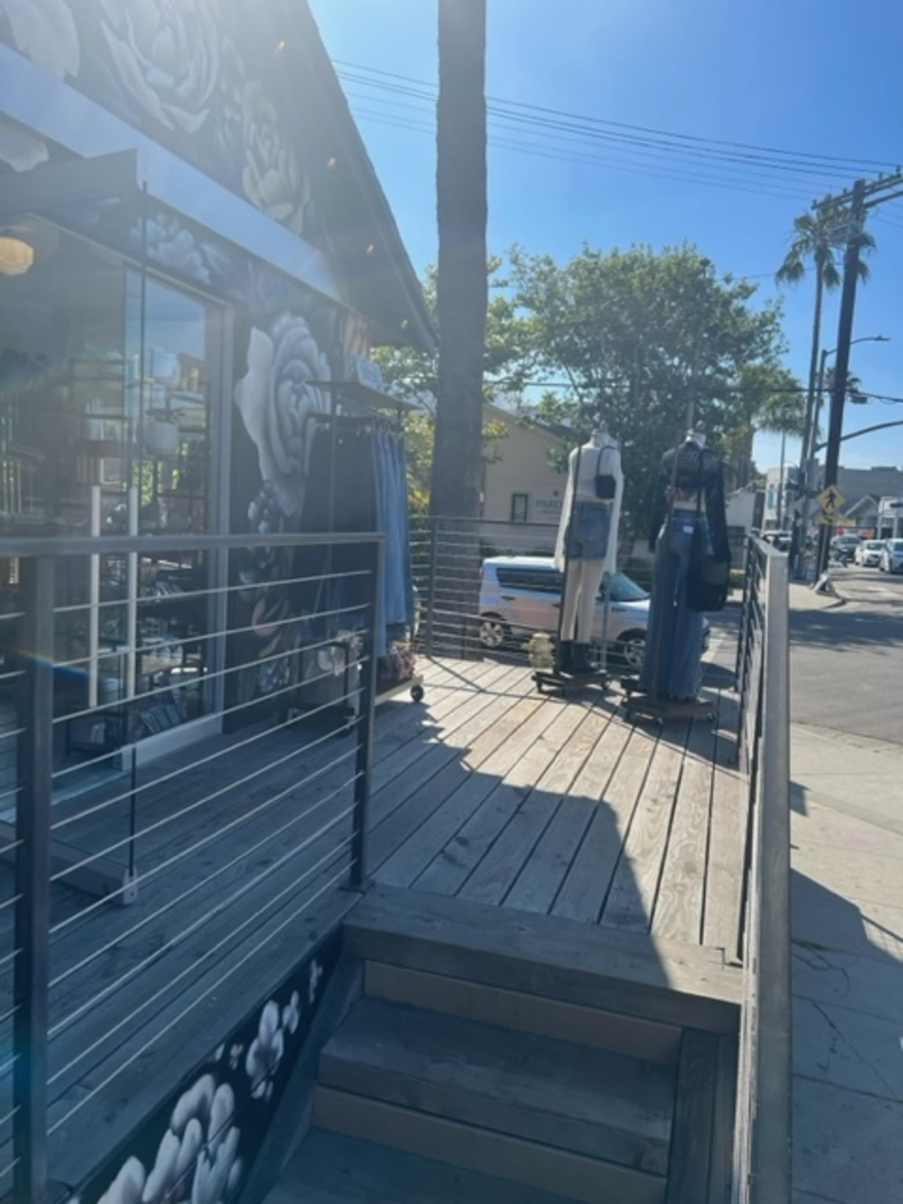 The image shows a wooden deck leading to a shop entrance with mannequins displaying clothing, surrounded by palm trees and a clear blue sky.