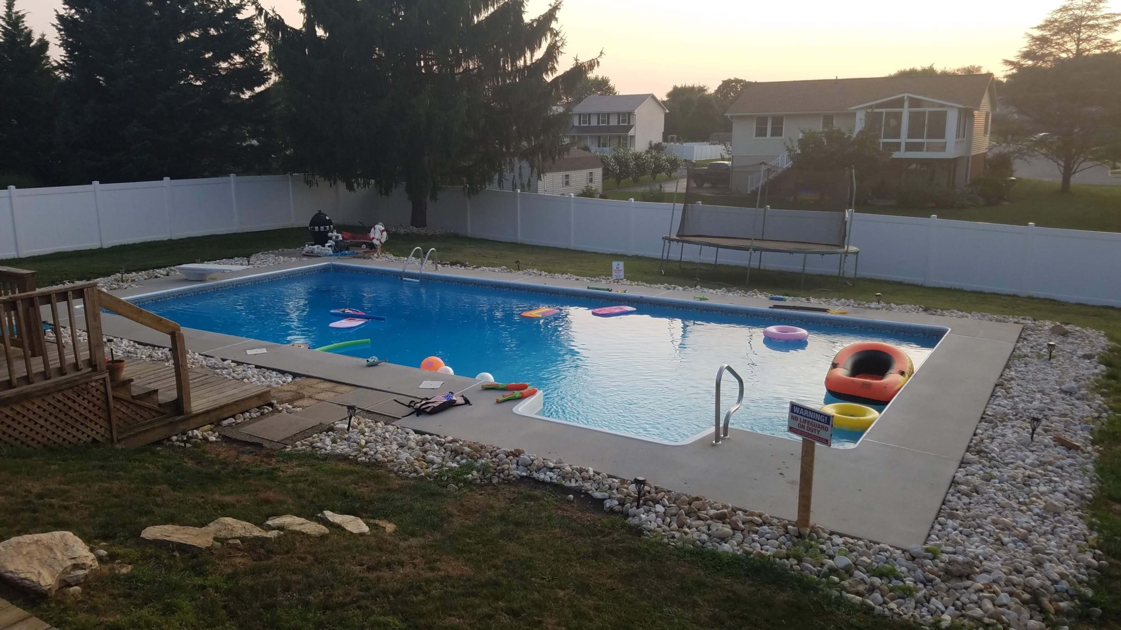 A backyard swimming pool surrounded by colorful inflatable floaties, a wooden deck, and a trampoline in a fenced yard at sunset.