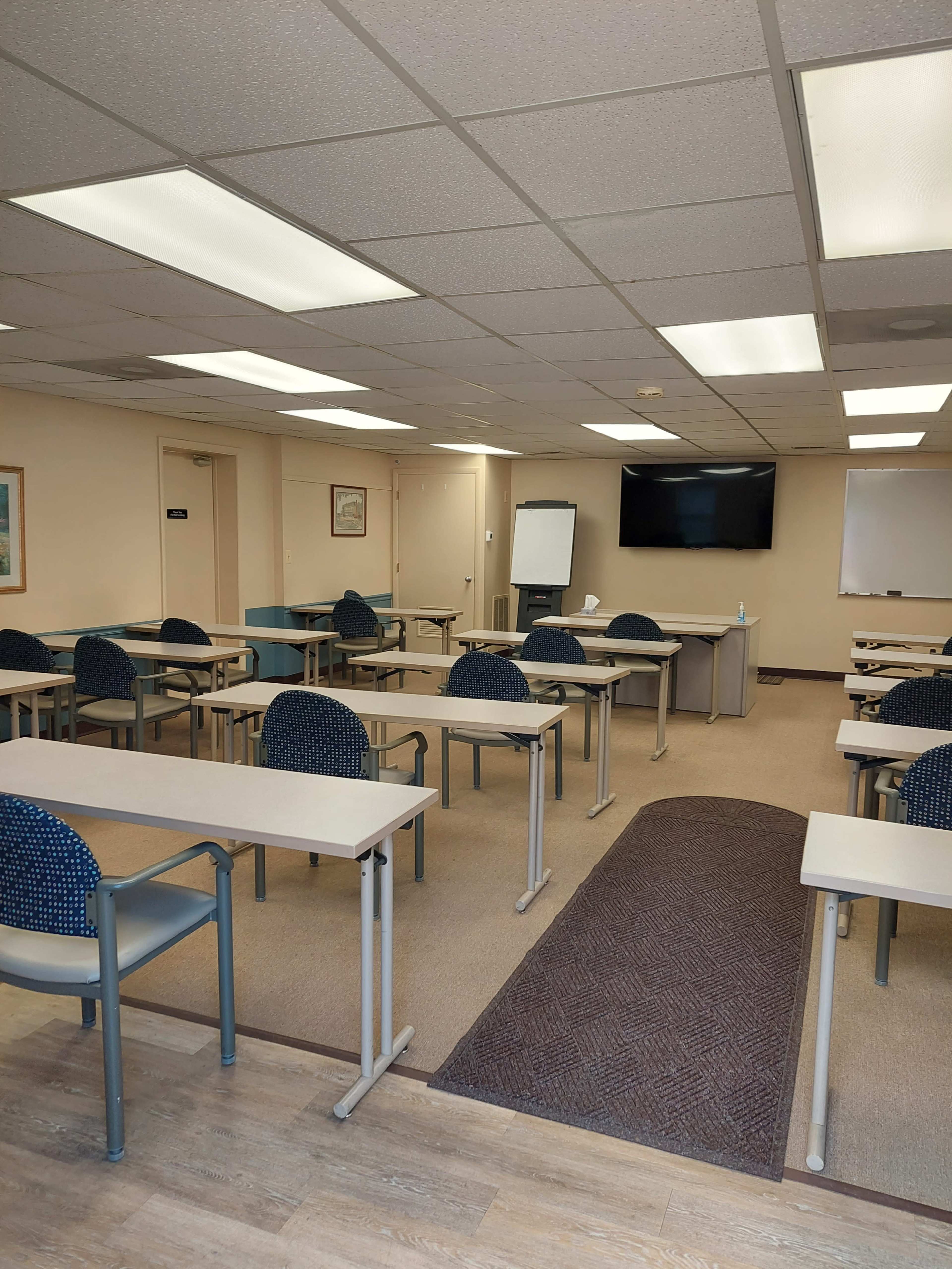 A classroom with rows of empty desks and chairs, a whiteboard, and a television mounted on the wall.