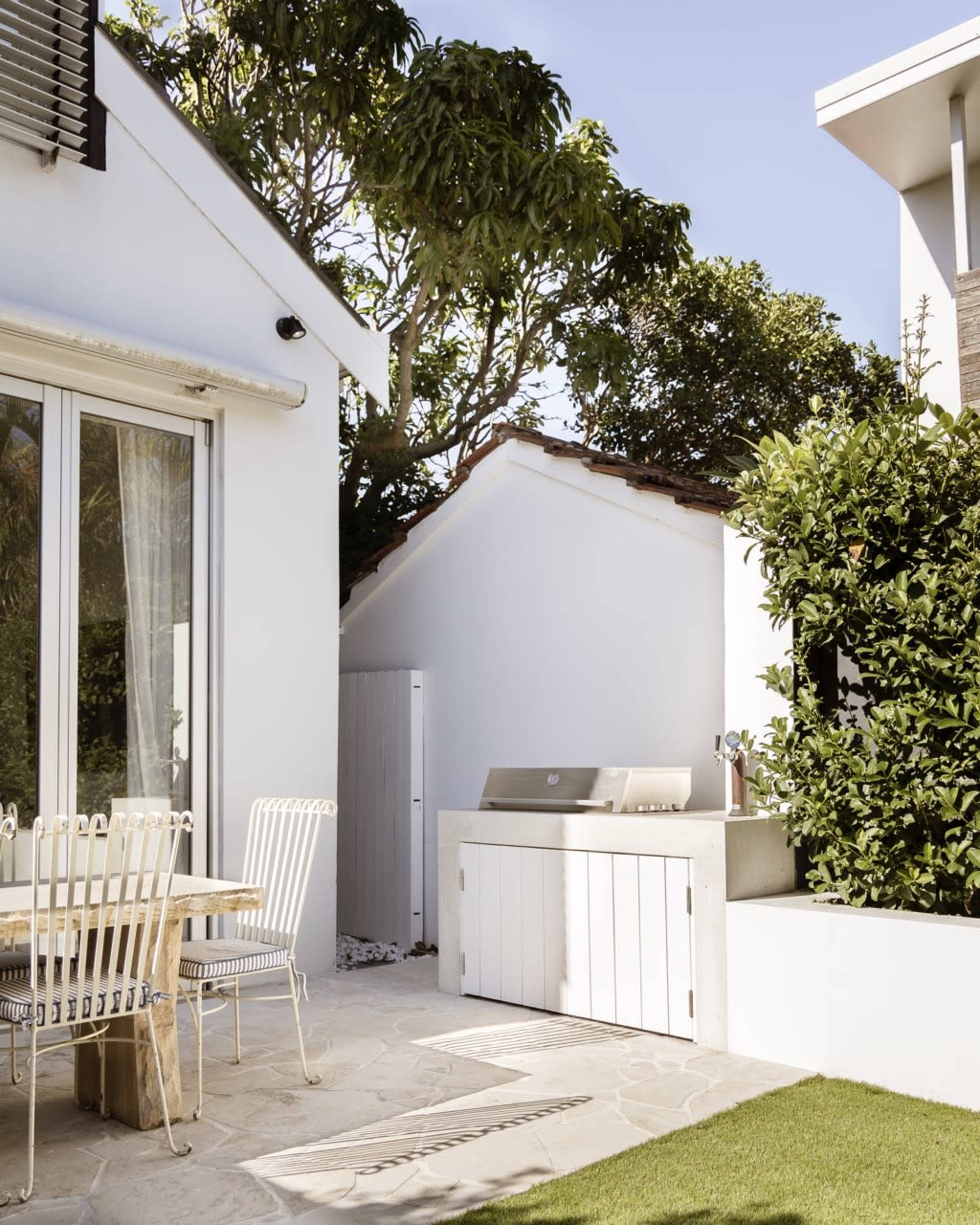 A modern outdoor space features a grill station adjacent to a white building, surrounded by greenery and a dining table with metal chairs.