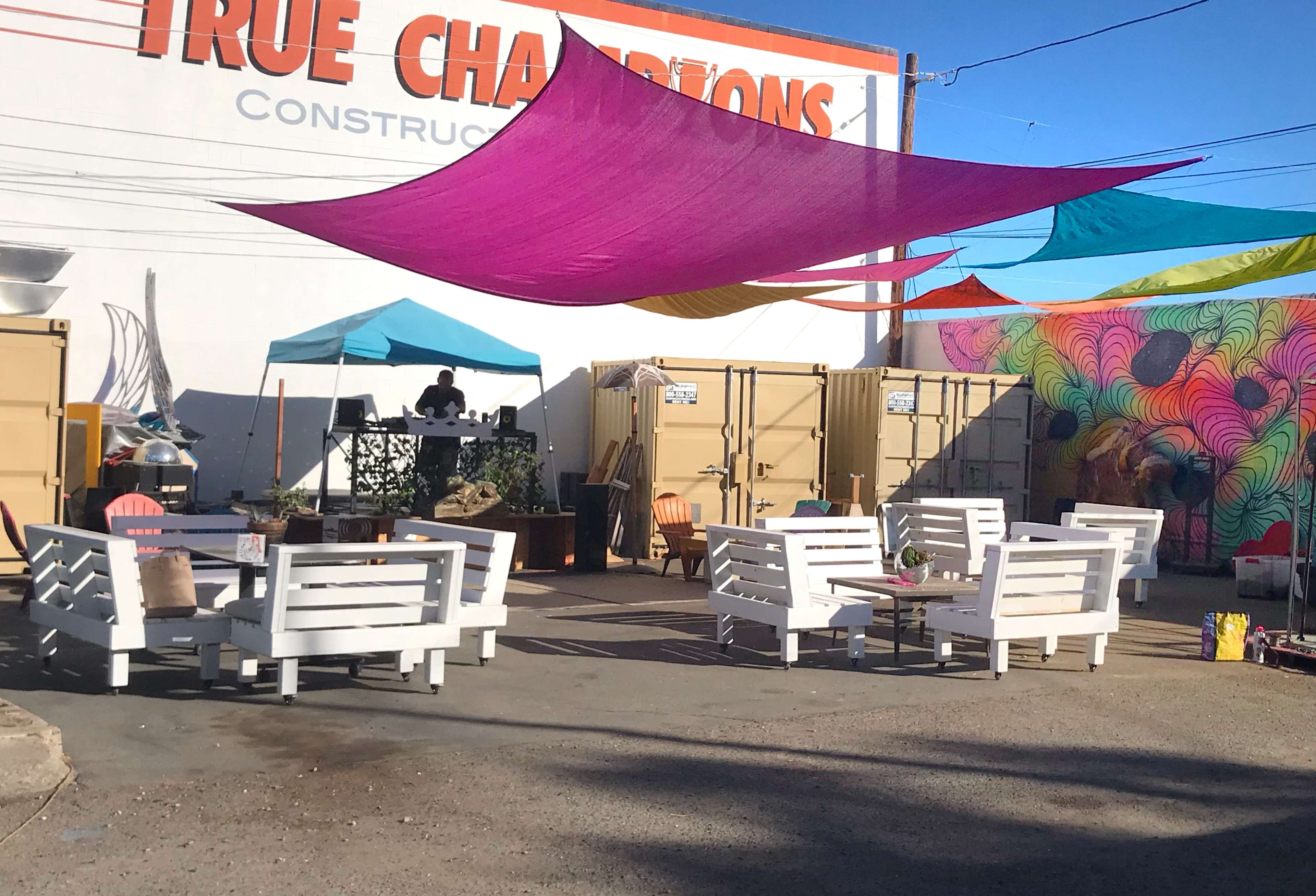 The scene features a colorful outdoor seating area with white furniture, various canopies, and a mural on a wall behind shipping containers.