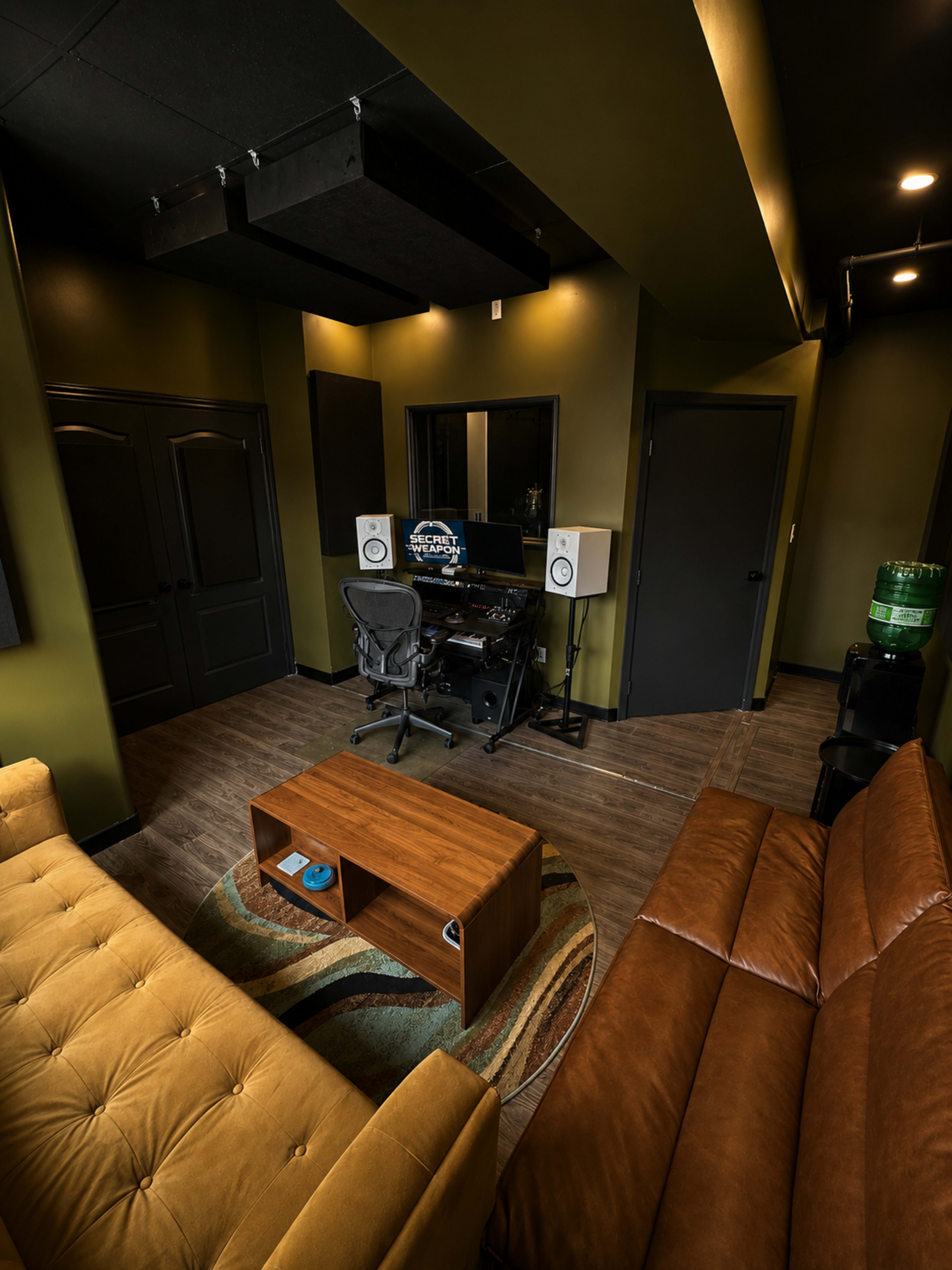 The image depicts a modern recording studio with dark green walls, a black desk with speakers, and seating areas furnished with a tan couch and a wooden coffee table.