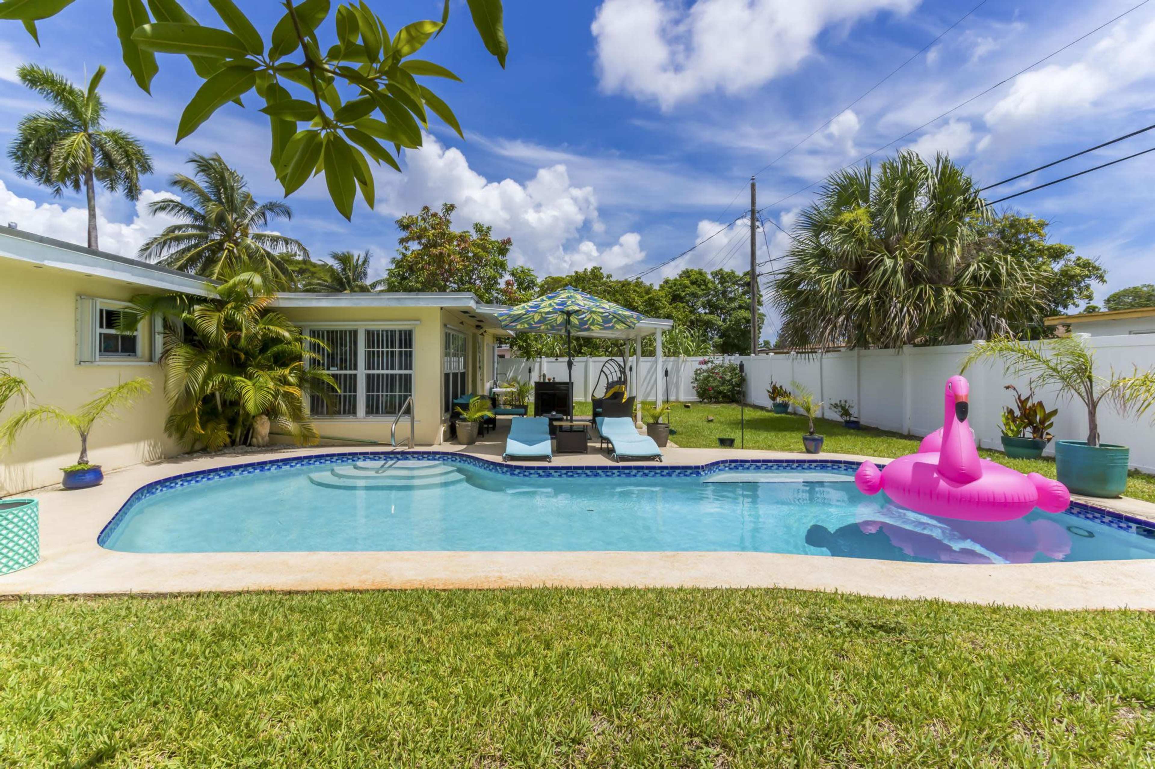 The image shows a swimming pool surrounded by greenery, lounge chairs, and a pink inflatable flamingo, with a clear blue sky above.
