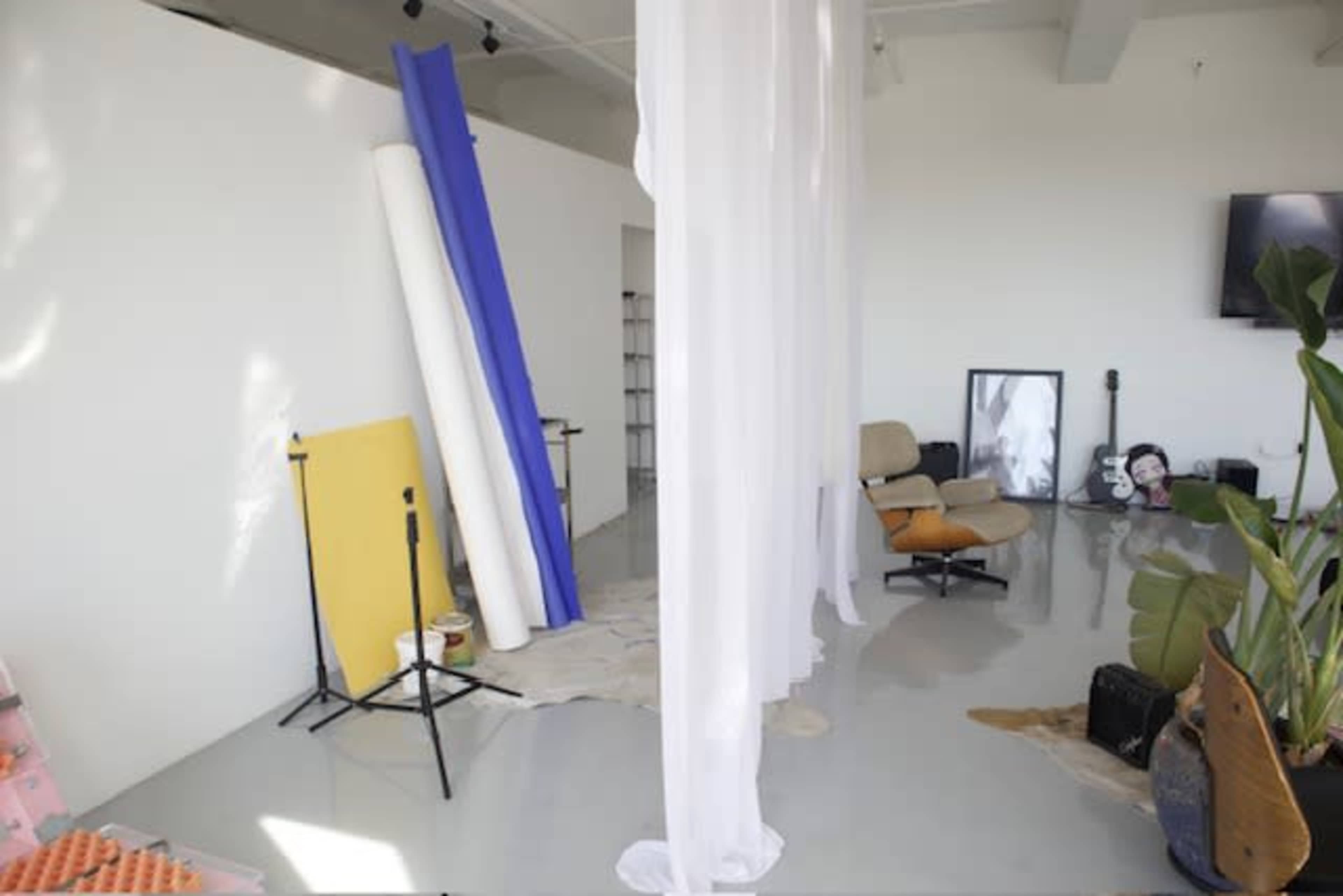 A spacious studio with white walls, featuring large rolls of colored paper, a chair, and various art supplies scattered on the floor.