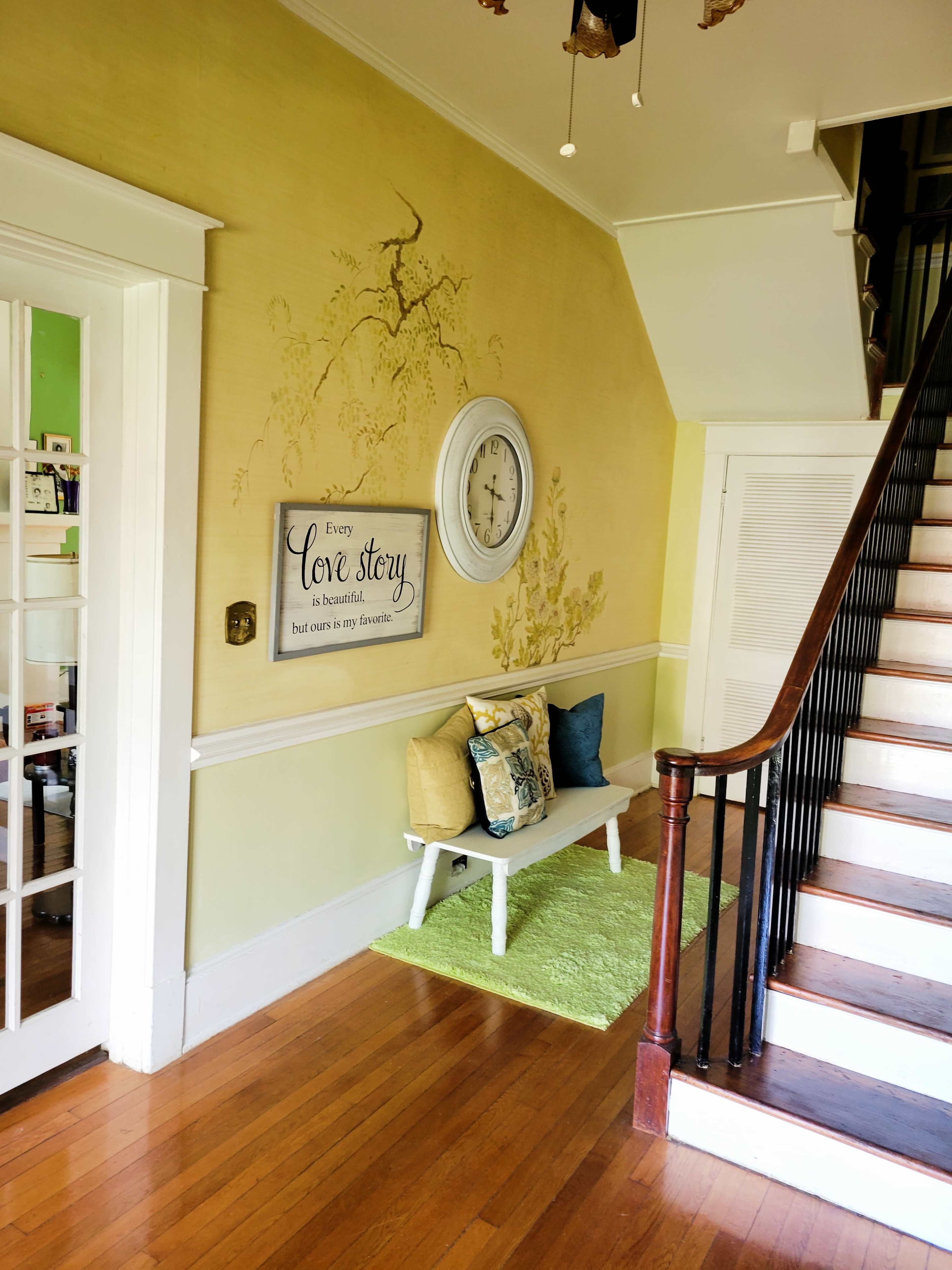 A hallway with a wooden staircase on the right, a clock and a decorative sign on the wall, and a bench with pillows on a green rug.