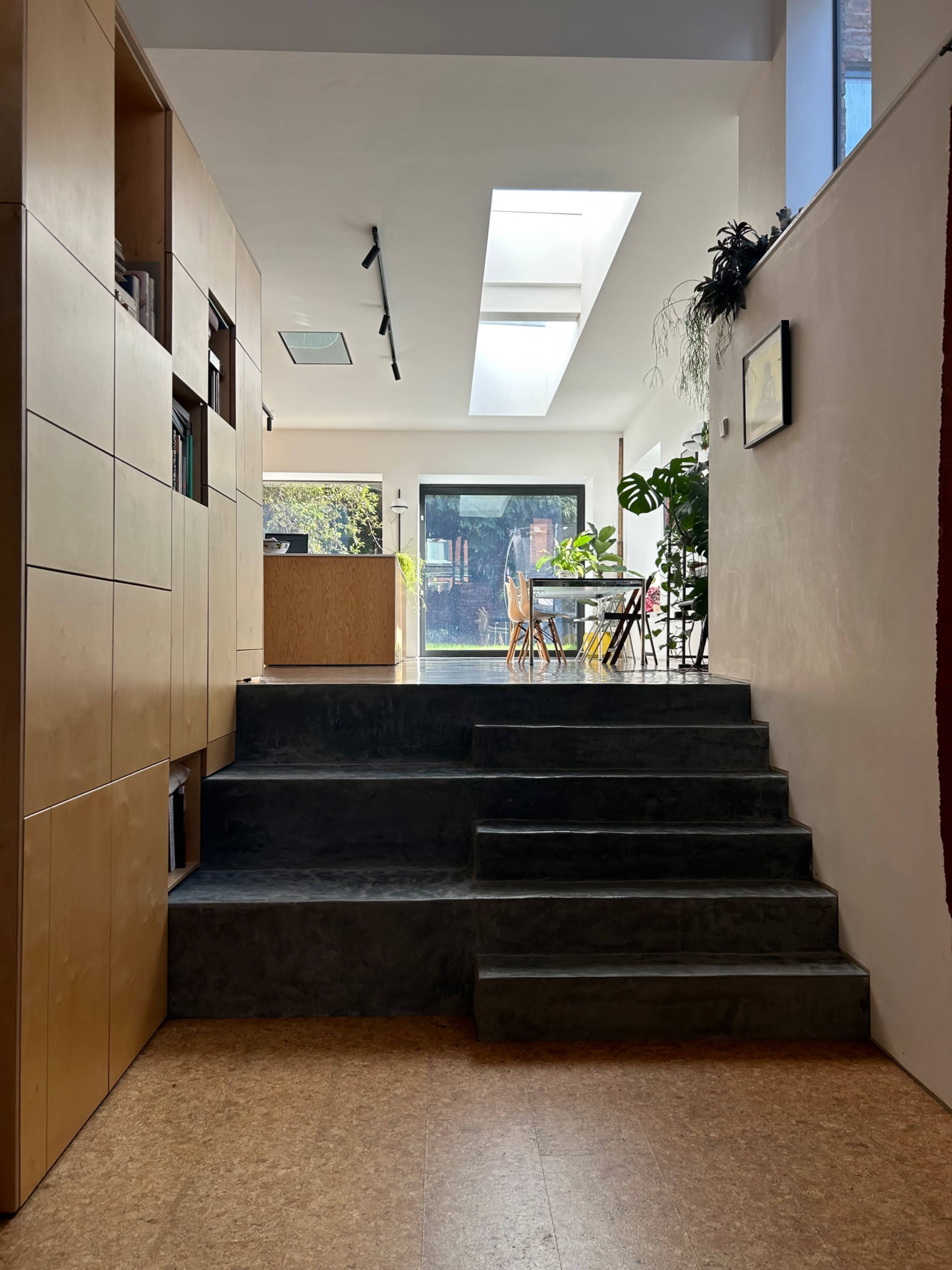 A modern interior space featuring wooden shelving, a set of stairs leading to a kitchen area, and large windows allowing natural light to fill the room.