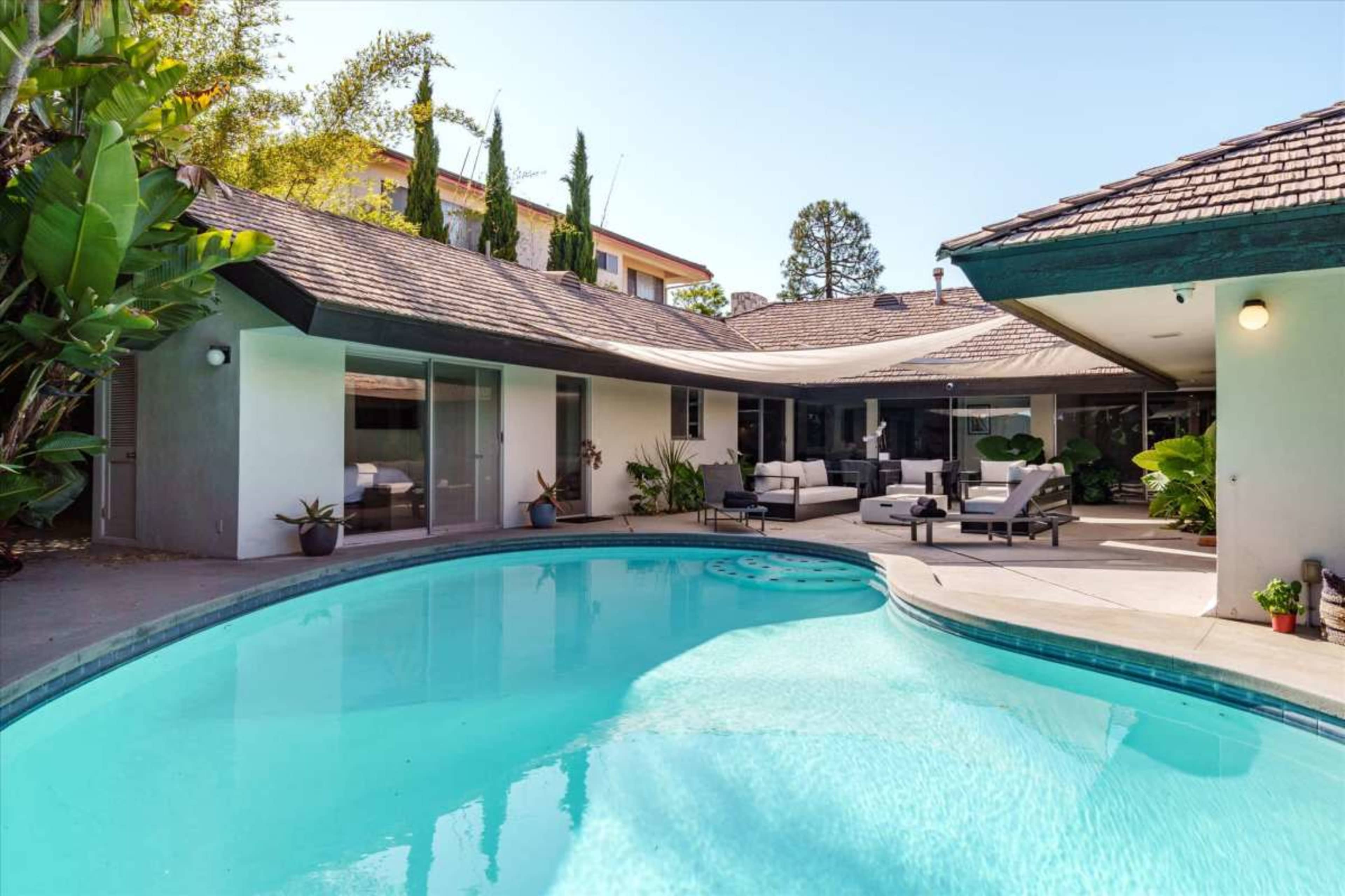 A swimming pool is situated in the foreground, surrounded by a patio area with outdoor furniture and tropical plants near a modern home.