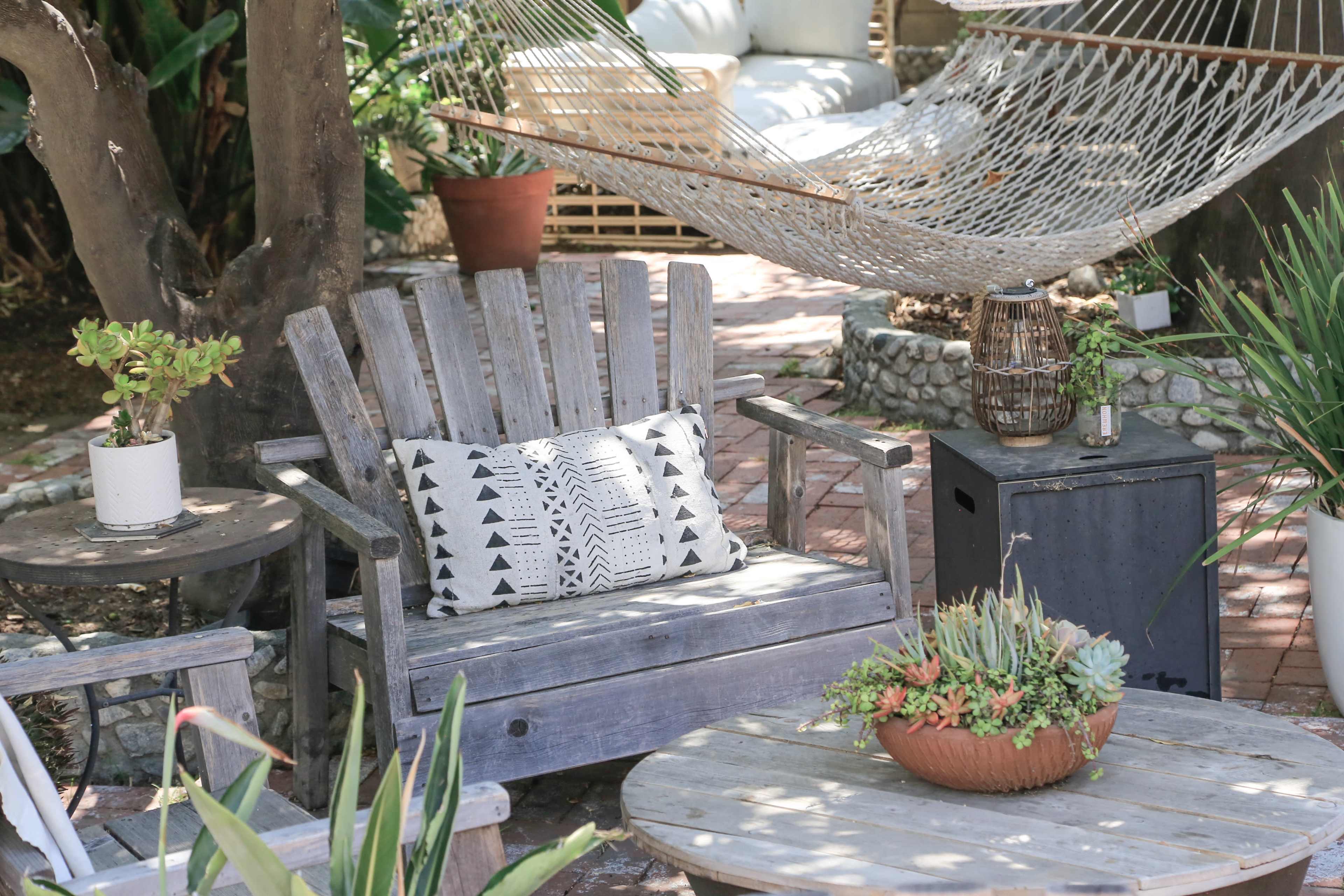 A wooden chair with a patterned pillow sits beside a rustic table in a garden area featuring a hammock and various plants.