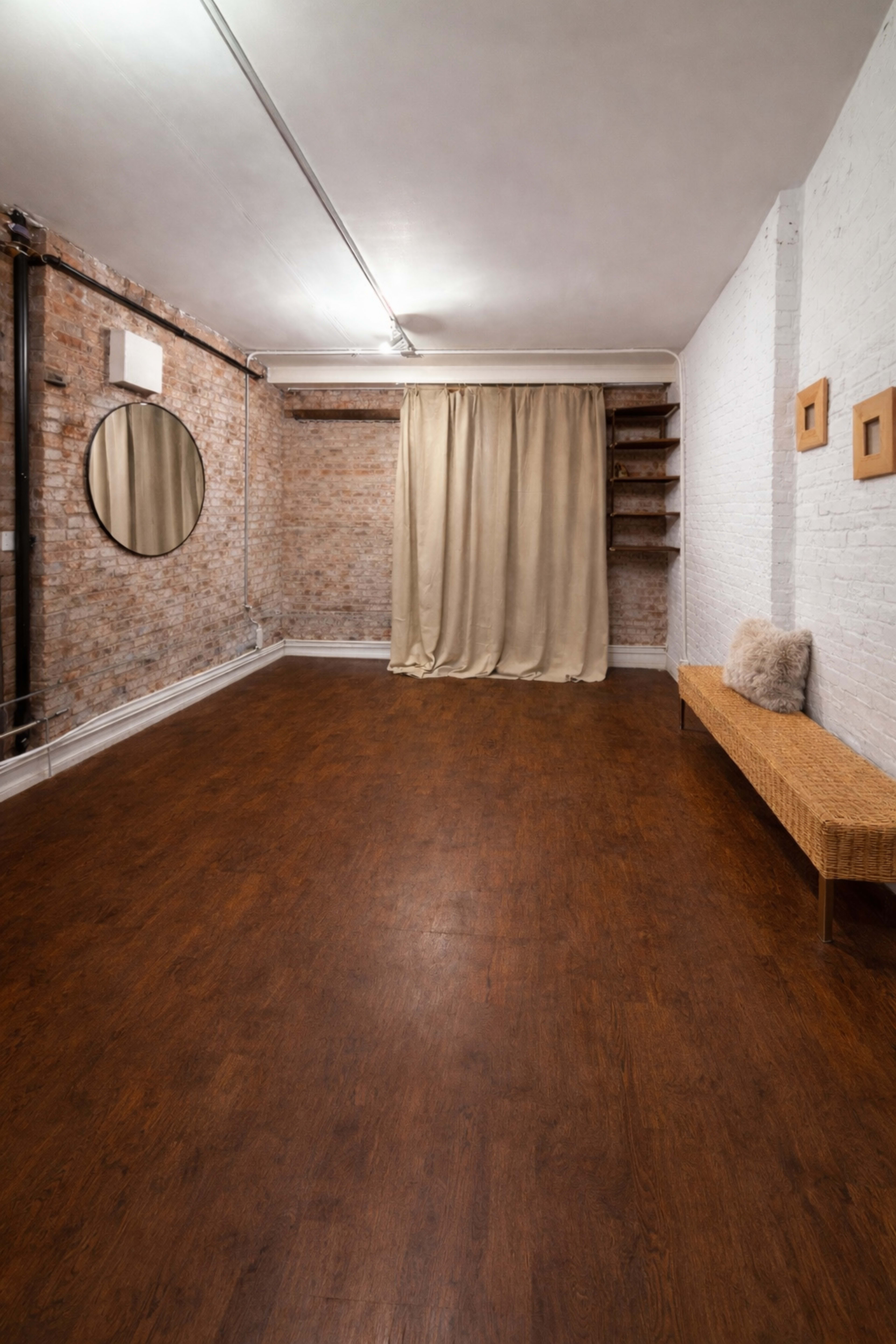 The image shows a spacious, empty room with exposed brick walls, a round mirror, a curtain, wooden flooring, and minimal furnishings.