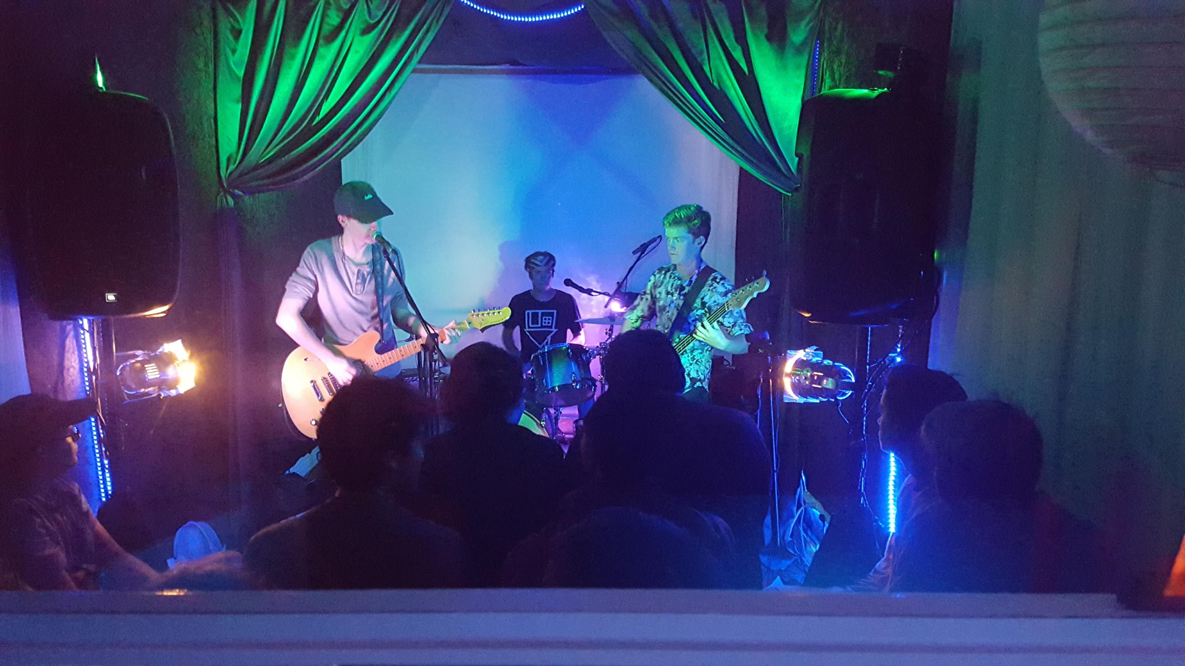 Two musicians play guitar and drums on a small stage with colorful lighting in front of an audience.