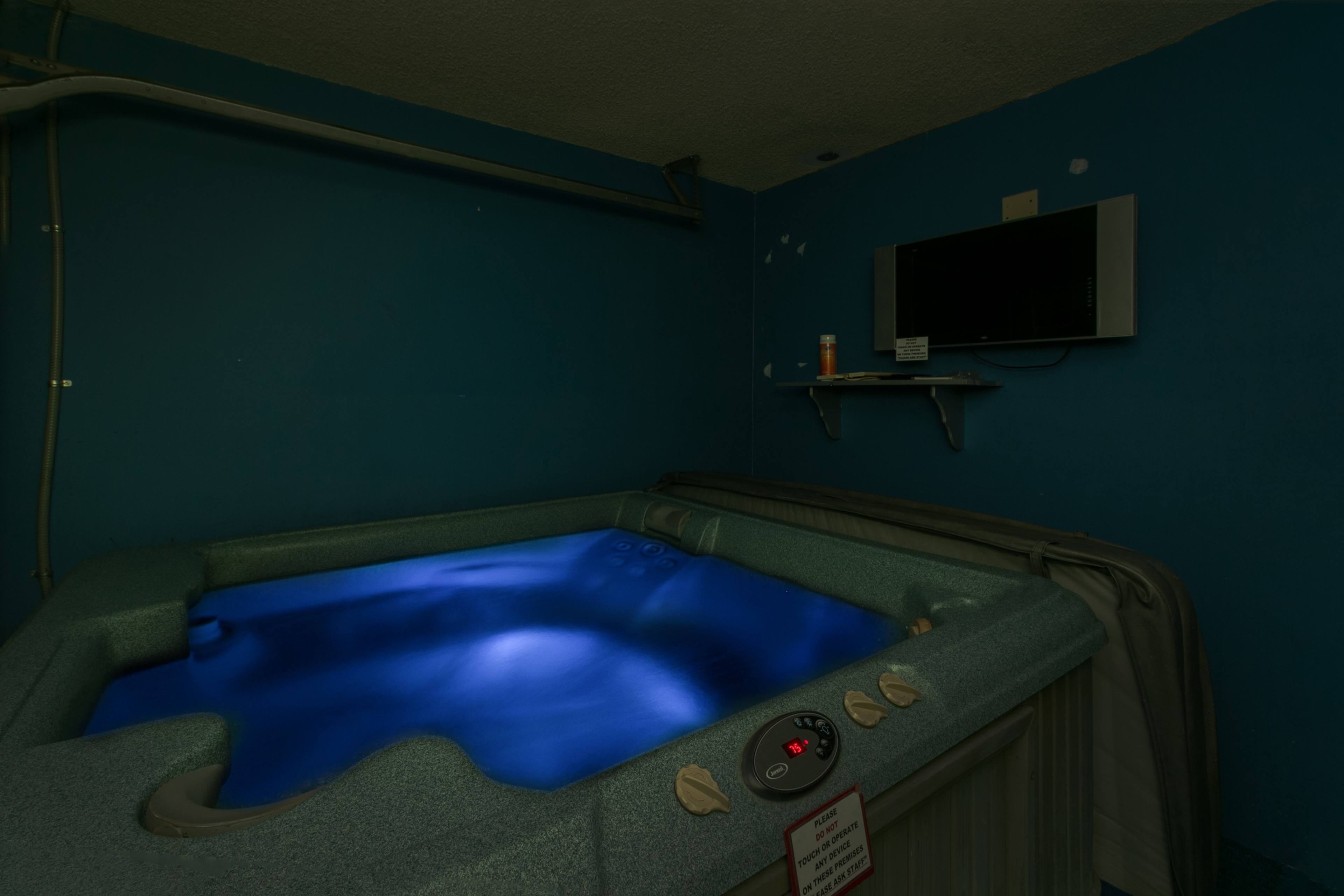 A hot tub with blue lighting is positioned in a room with a blue wall and a mounted television.