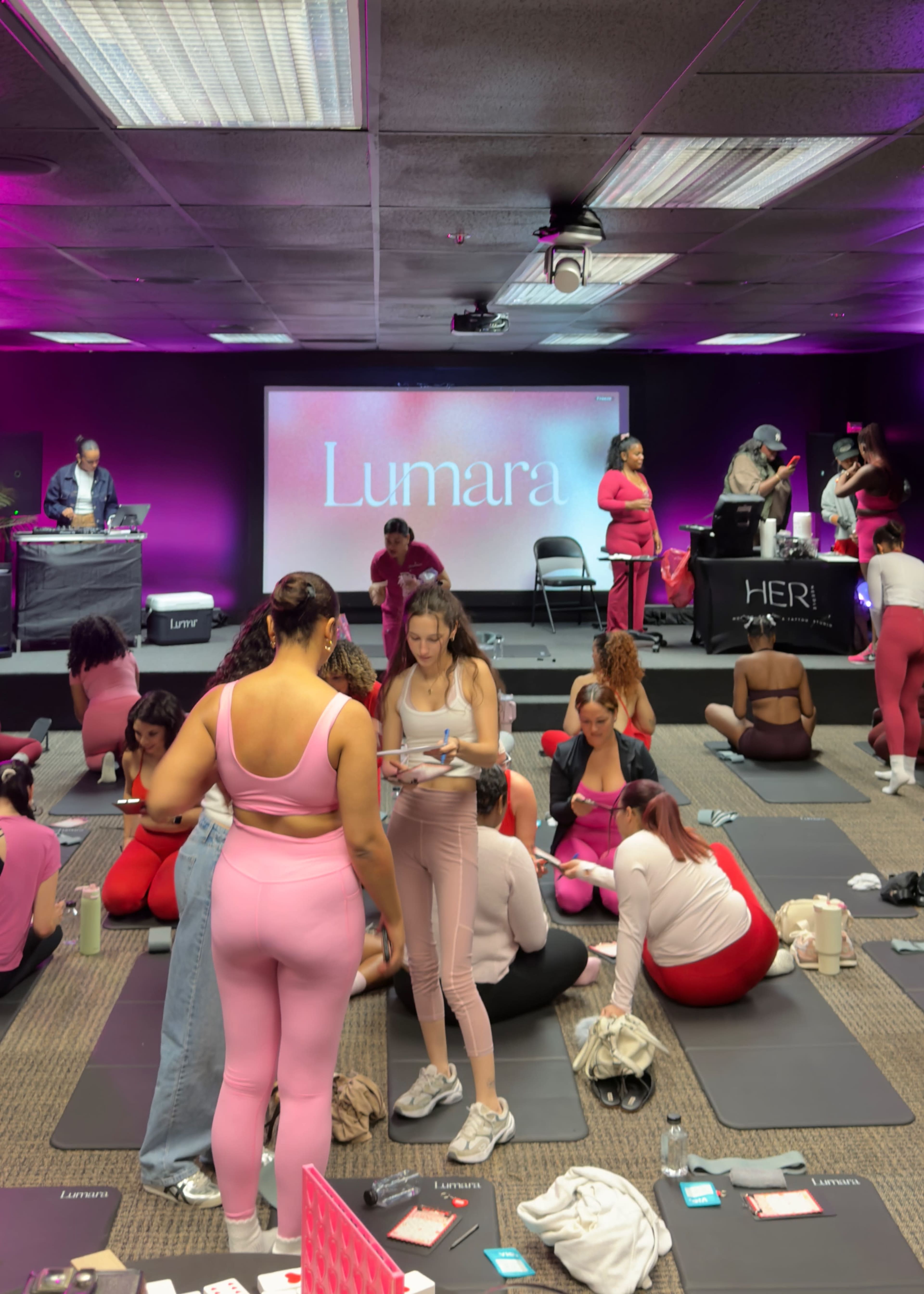 The image shows a fitness event with participants dressed in pink and red workout attire, arranged in a room featuring a large screen displaying the word "Lumara."