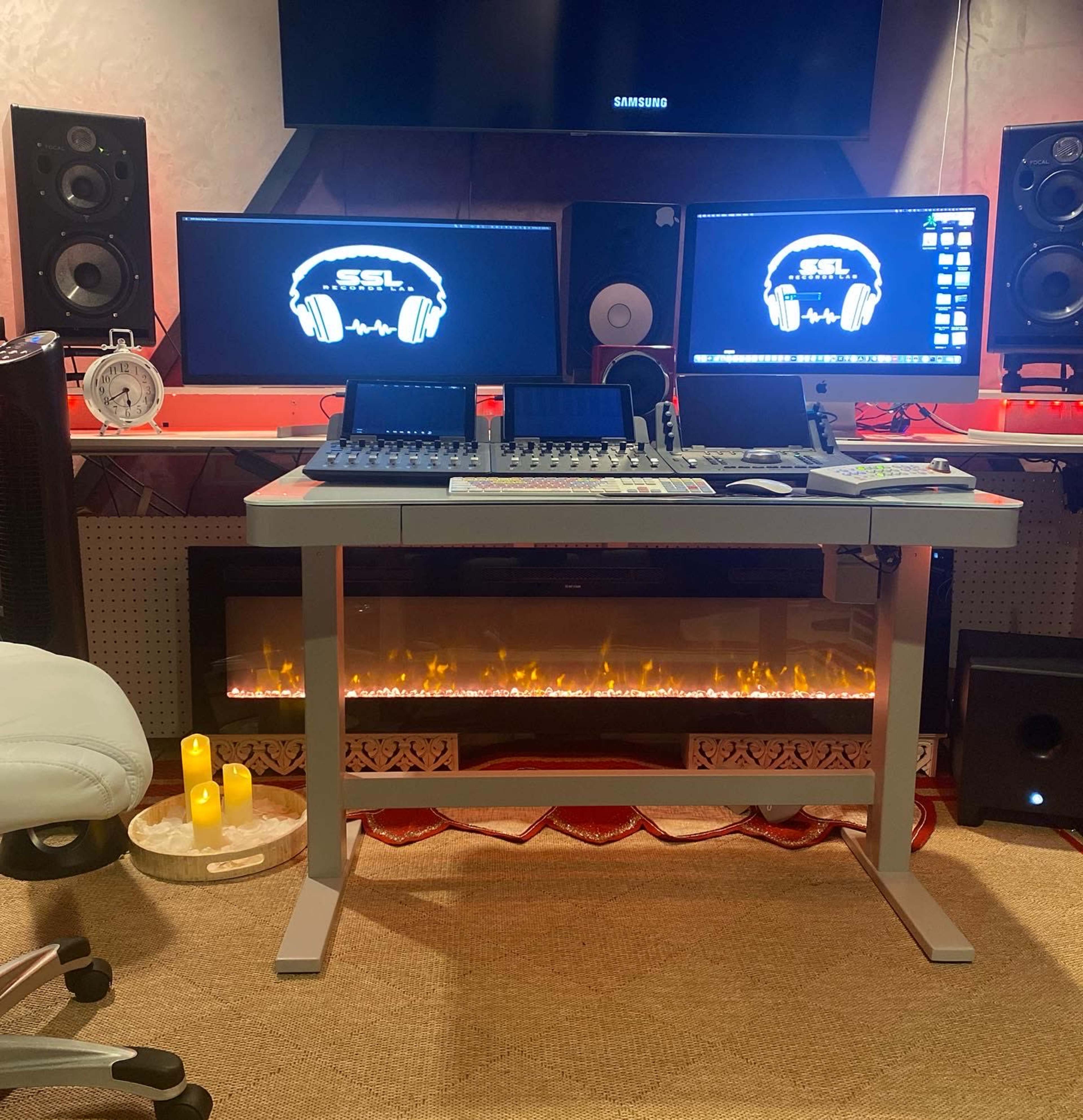 A home studio setup with two computer monitors displaying audio software, a digital audio mixer, and an electric fireplace underneath a modern desk.
