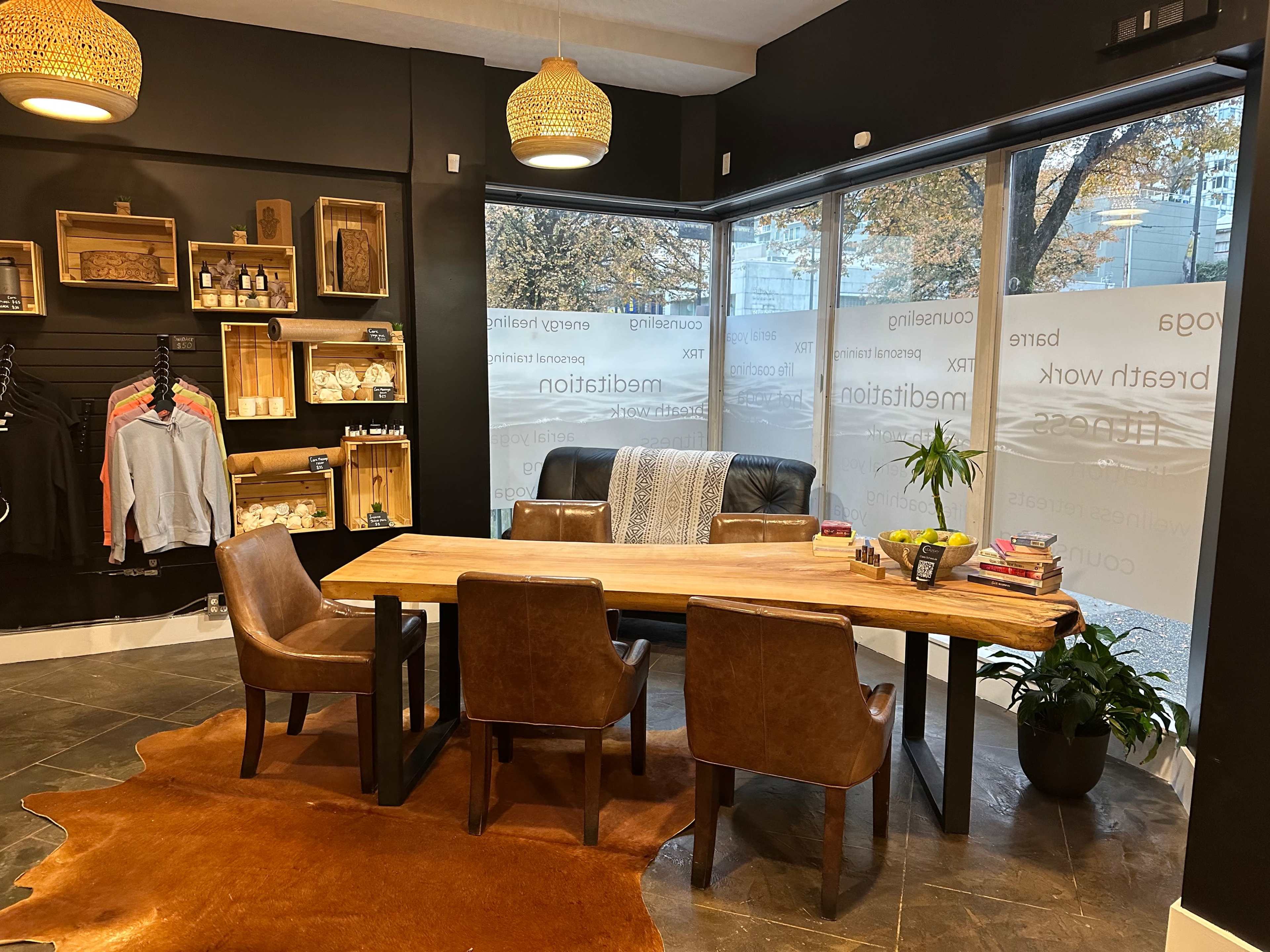 A modern office space features a wooden table surrounded by brown chairs, with large windows displaying a view of trees outside and shelves stocked with various items on the walls.