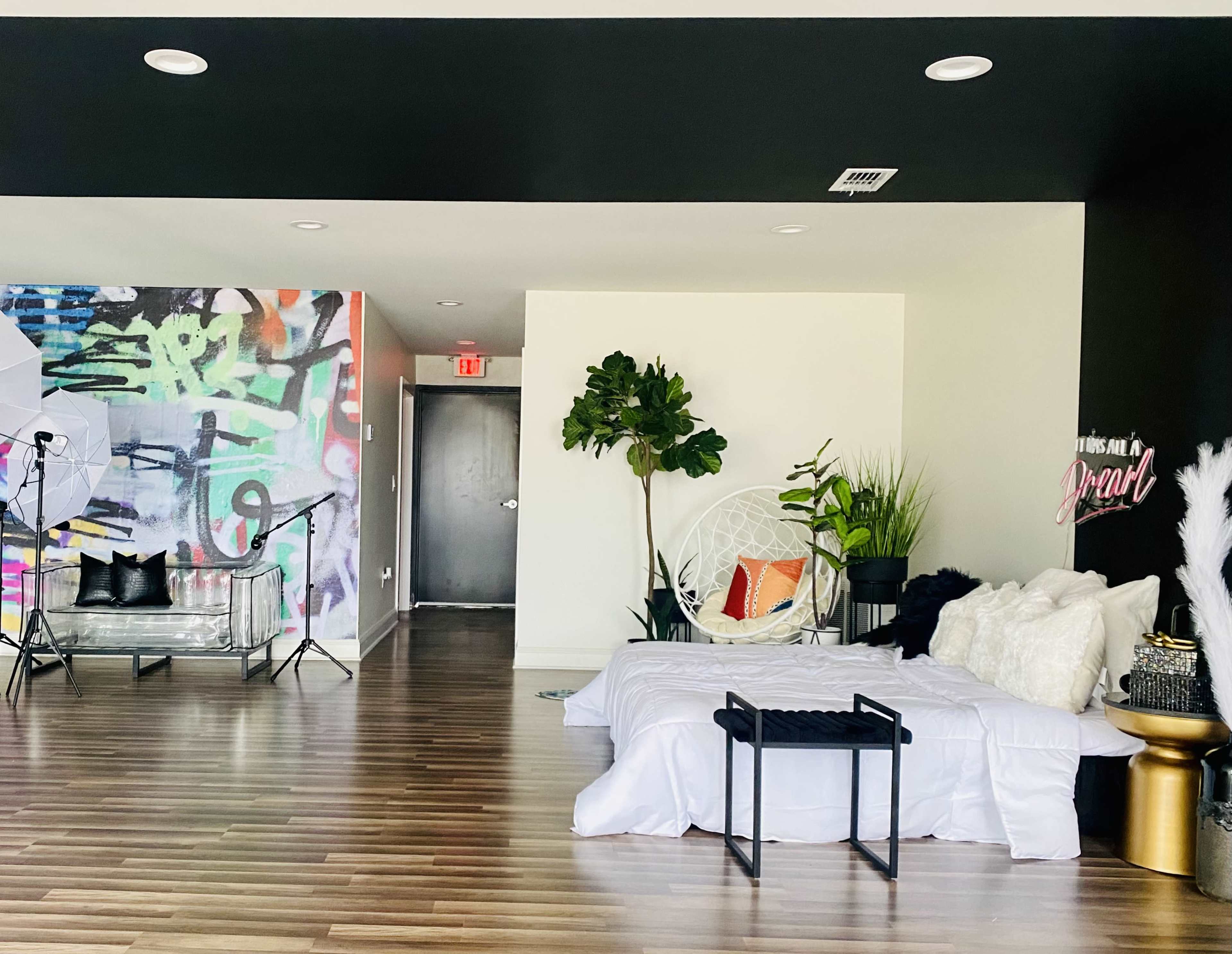 The image shows a spacious room with a bed covered in white linens, a decorative chair, and a mix of plants, along with a wall featuring colorful graffiti art.