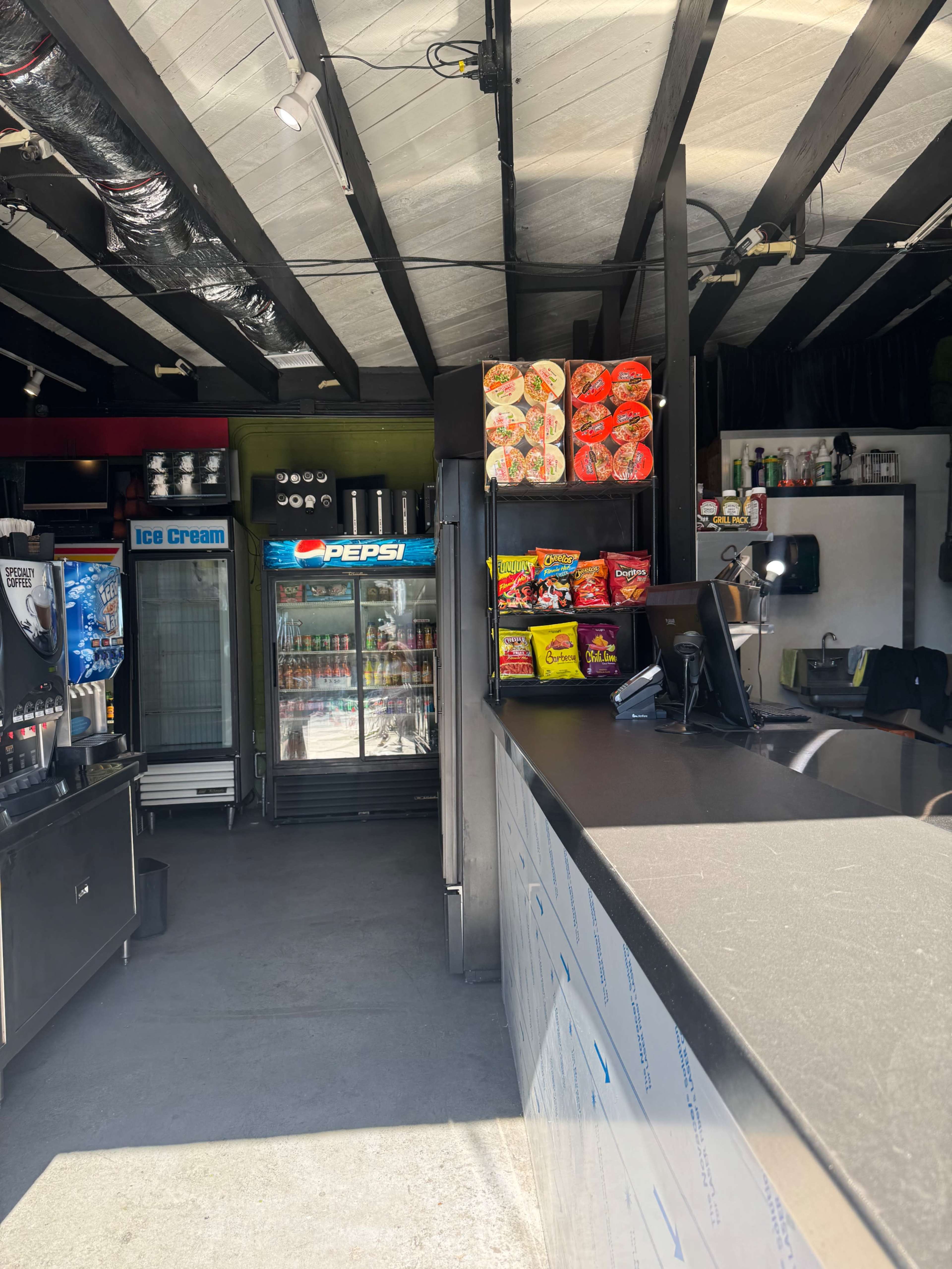 The image shows the interior of a small food and beverage kiosk featuring a counter, shelves stocked with snacks, and a glass refrigerator displaying drinks and ice cream.