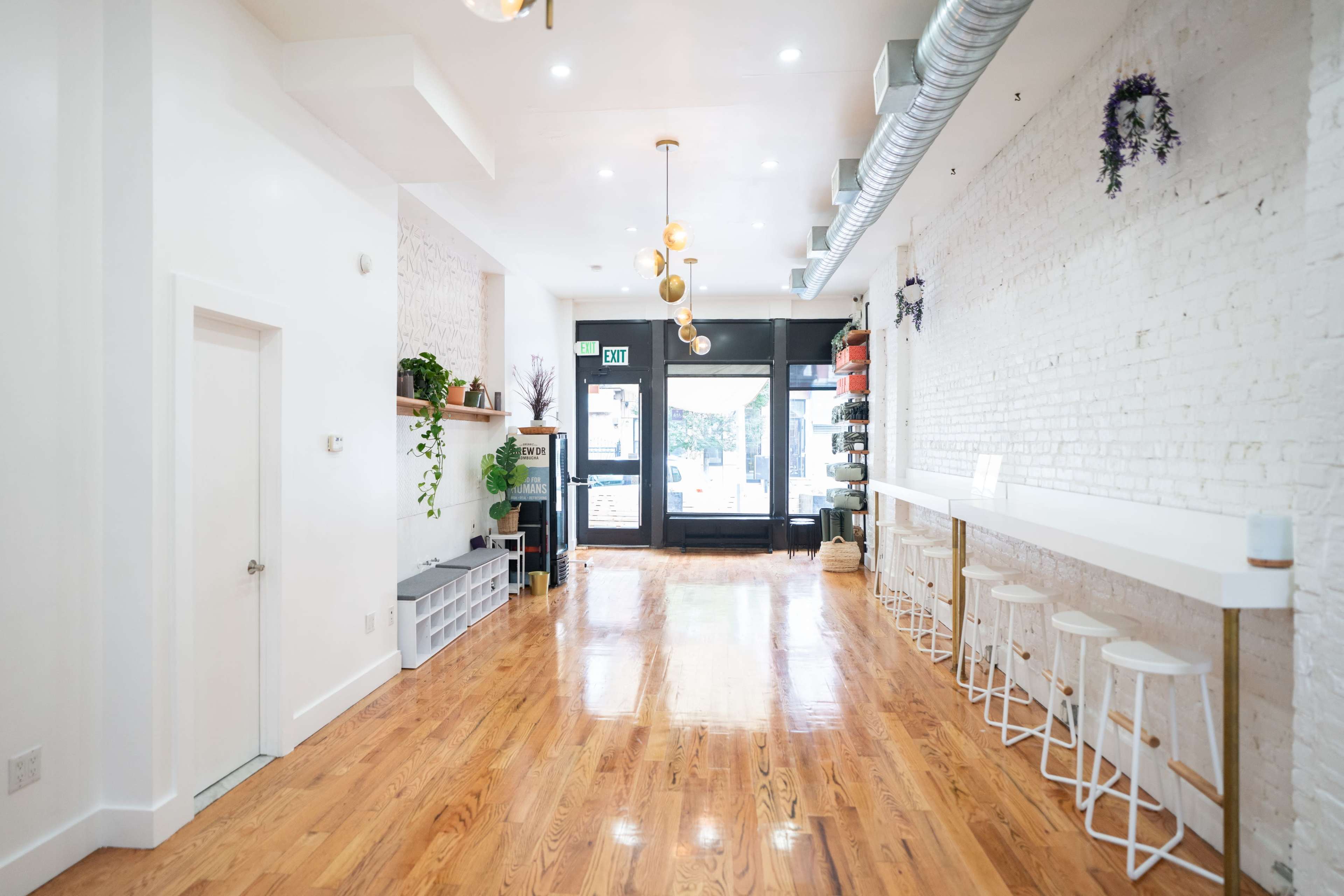 The interior features a spacious, minimalist design with wooden flooring, white walls, and a row of stools at a counter by large front windows.