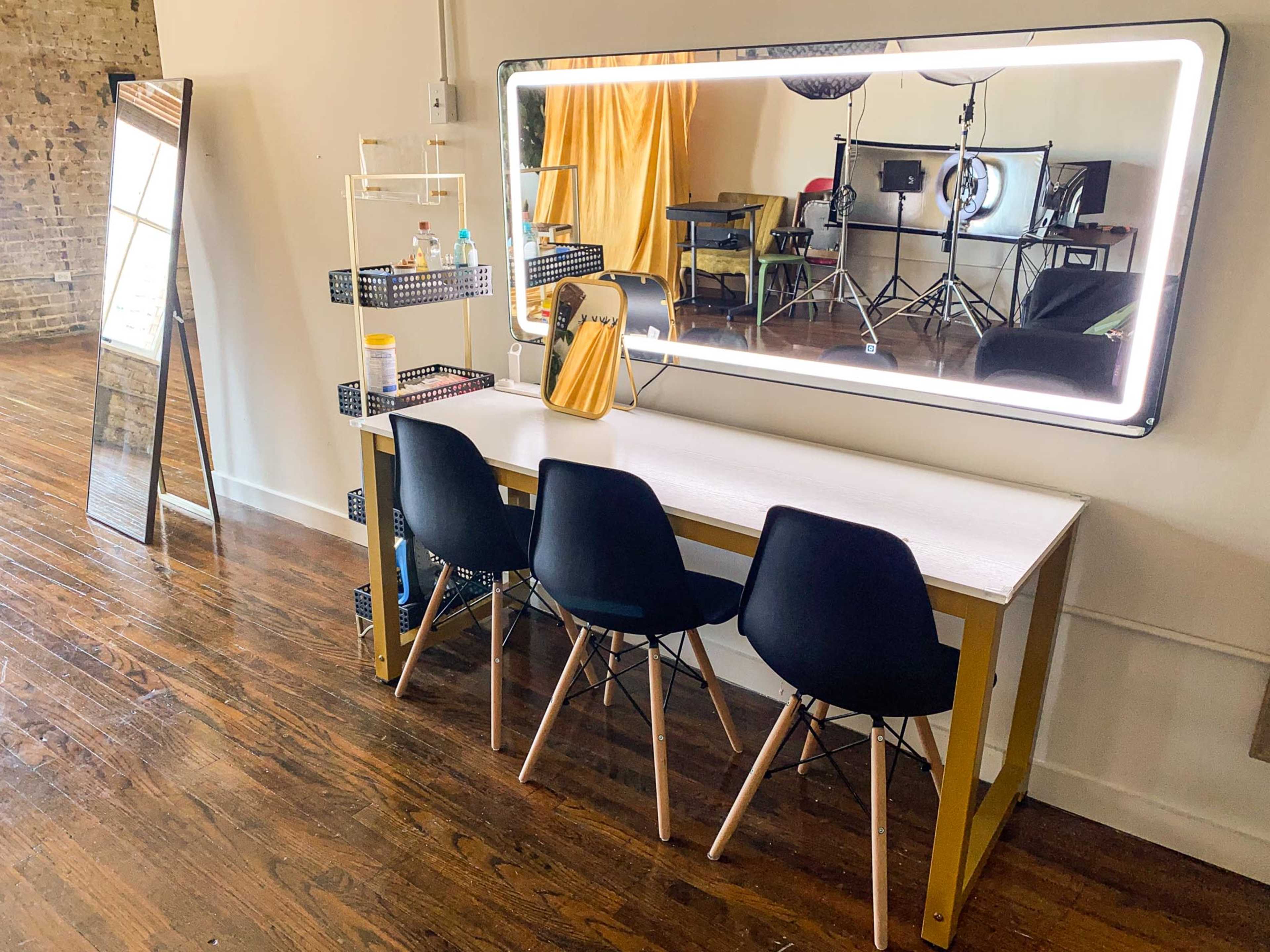 A vanity table with three black chairs, a large illuminated mirror, and a shelf with bottles, set in a spacious room with wooden floors and studio lighting equipment in the background.