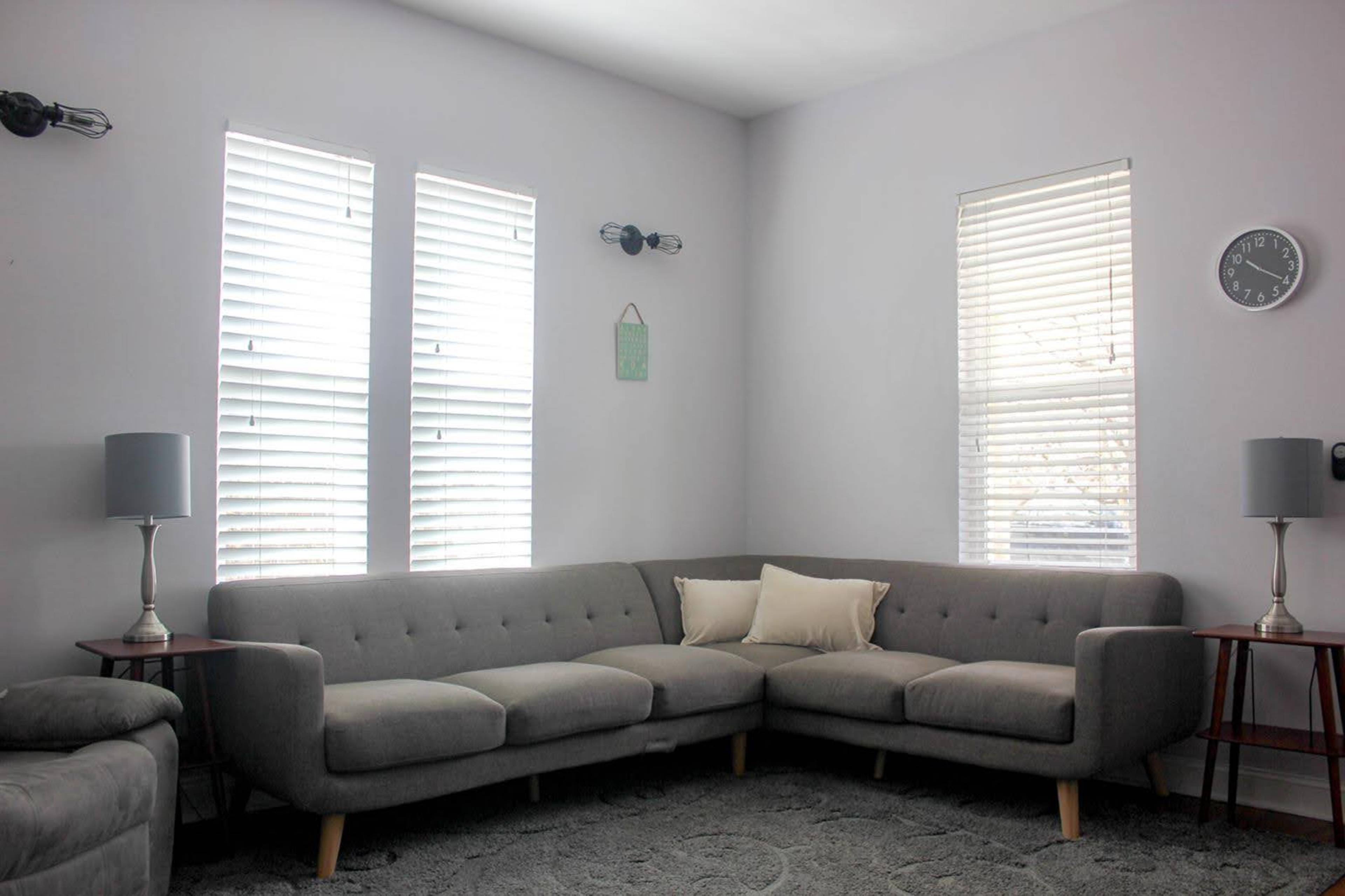 A modern living room features a gray sectional sofa, two lamps on wooden side tables, and bright windows with blinds.
