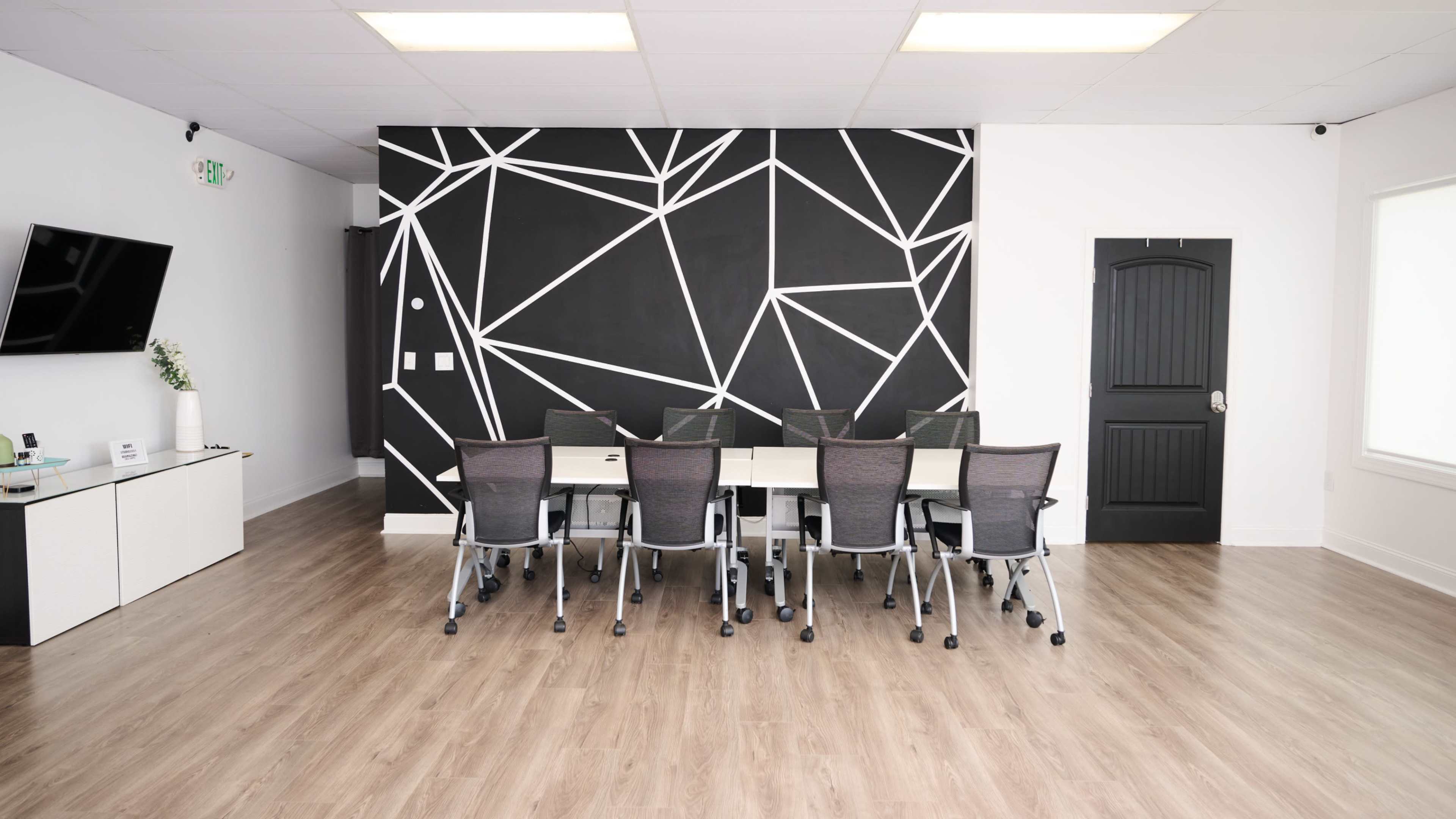 A conference room features a long table surrounded by rolling chairs, with a black geometric-patterned wall behind it.