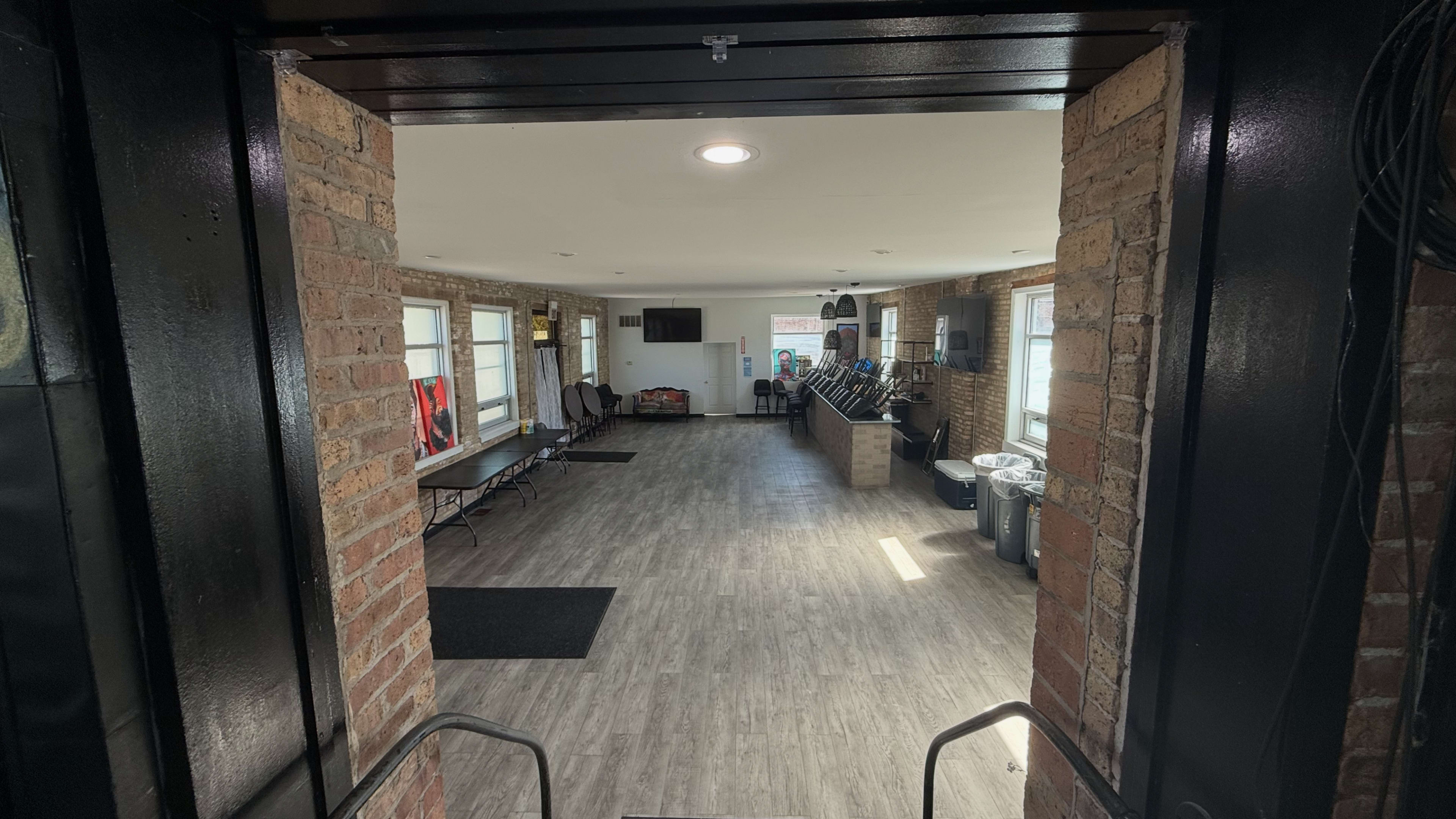 The image shows a spacious interior room with brick walls, large windows, and various seating and equipment along the sides.