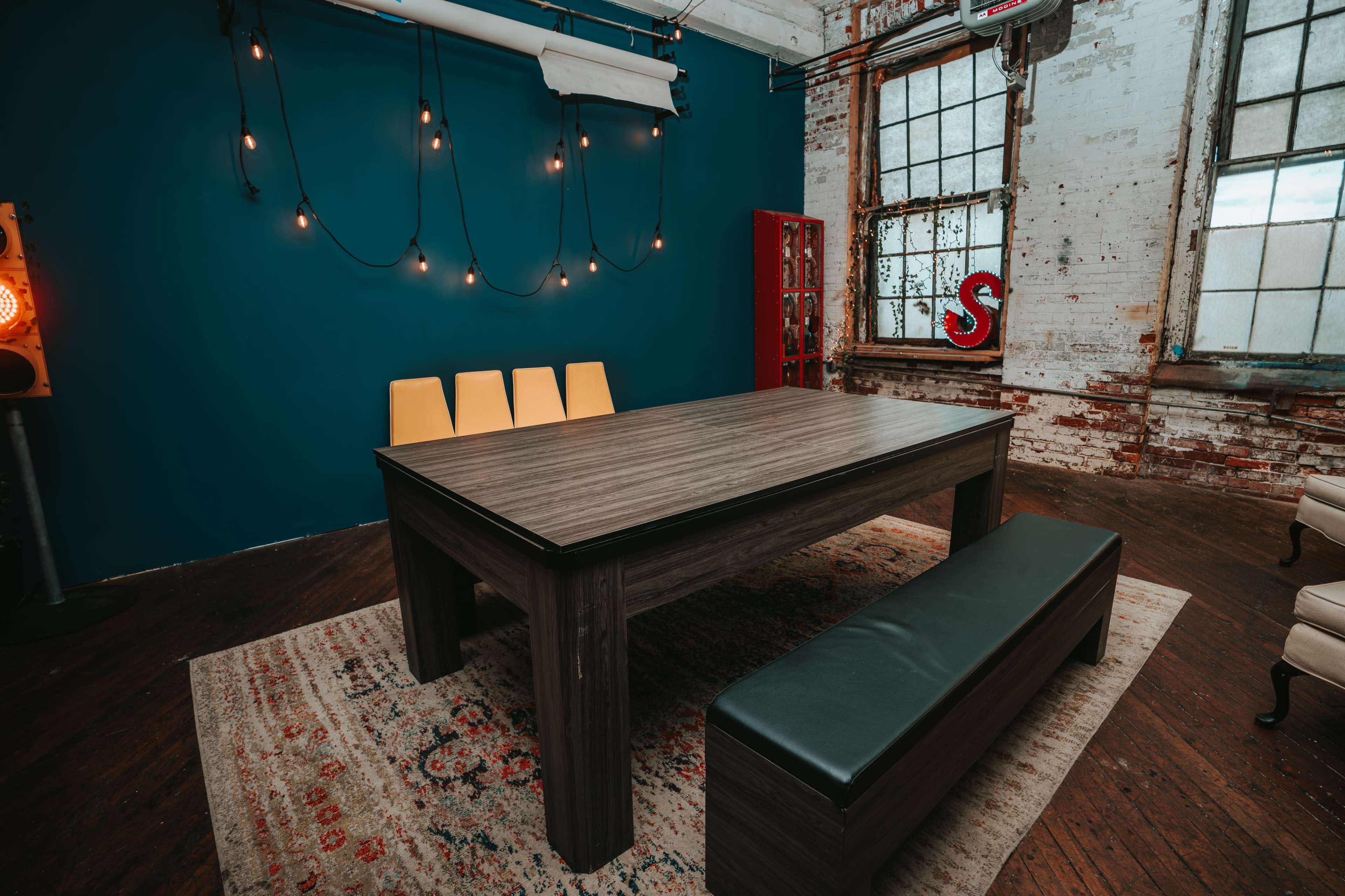 A large wooden table with a dark finish is placed on a patterned rug, surrounded by yellow chairs and a long bench in a room with a blue wall and industrial lighting.