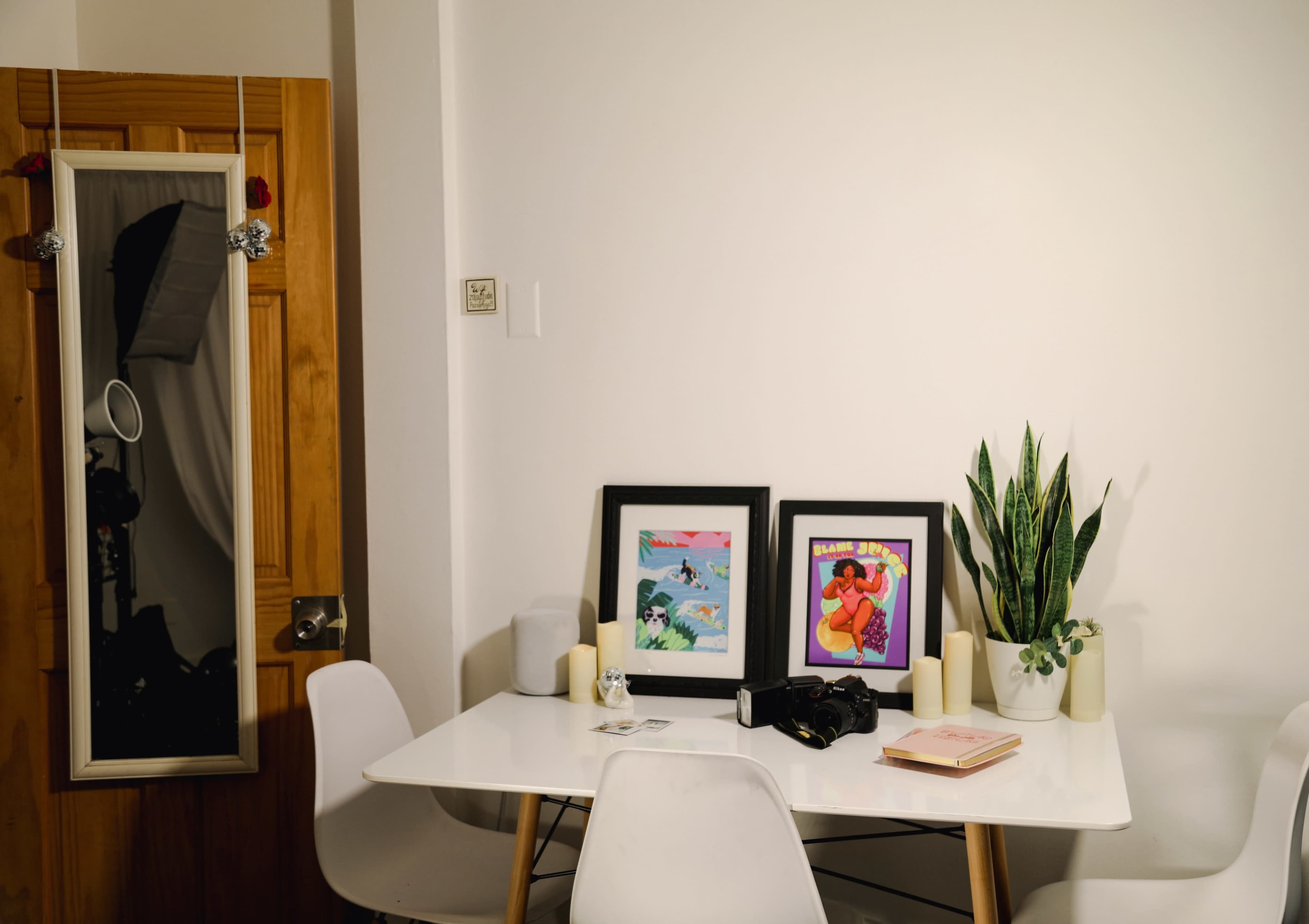 A white table is set up in a room with two framed pictures, candles, a plant, and a camera, while a mirror hangs on the wall beside a wooden door.