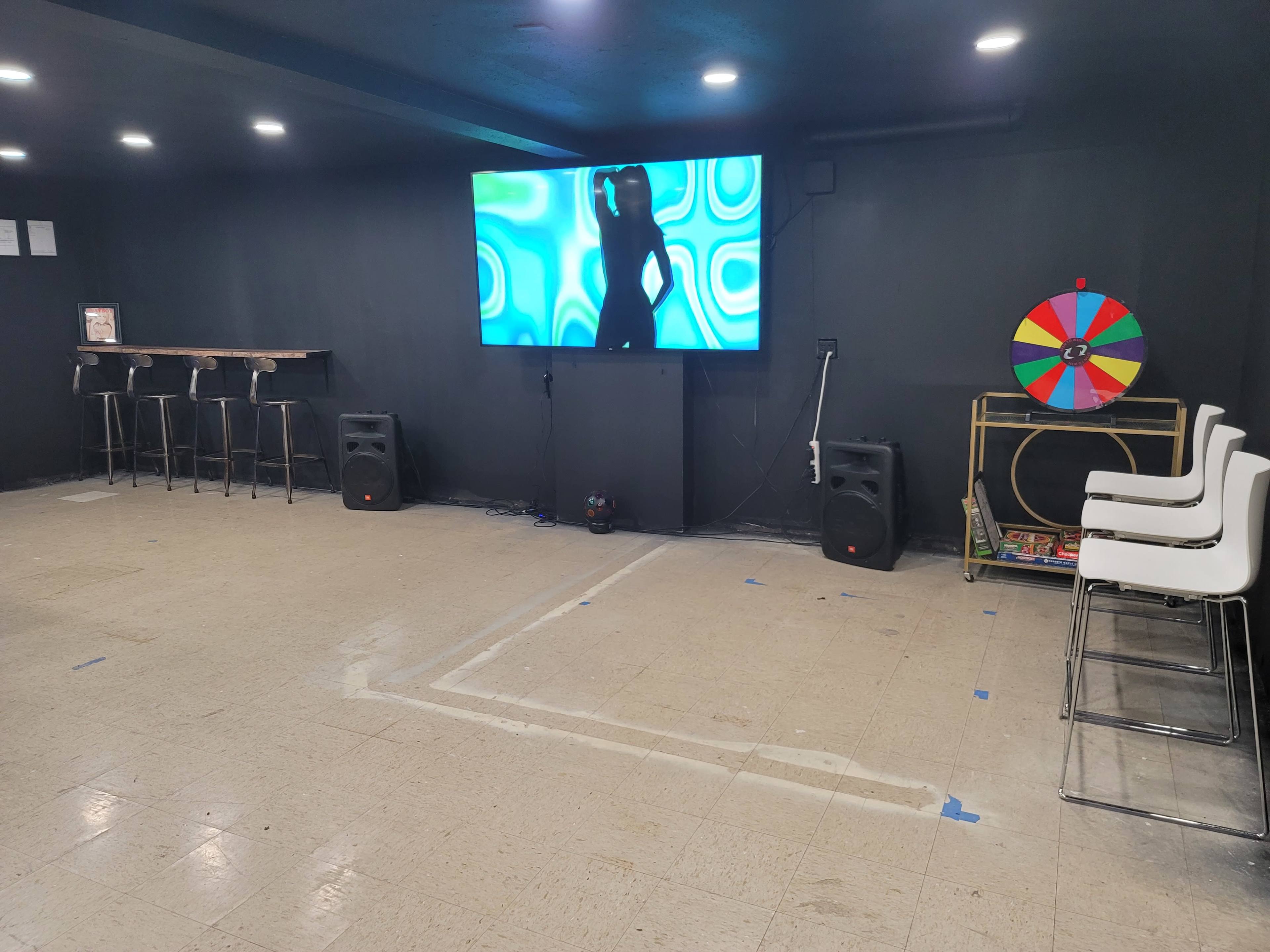 The room features a large screen displaying a silhouette, two speakers, a colorful spinning wheel on the wall, a bar table with stools, and a bare floor.