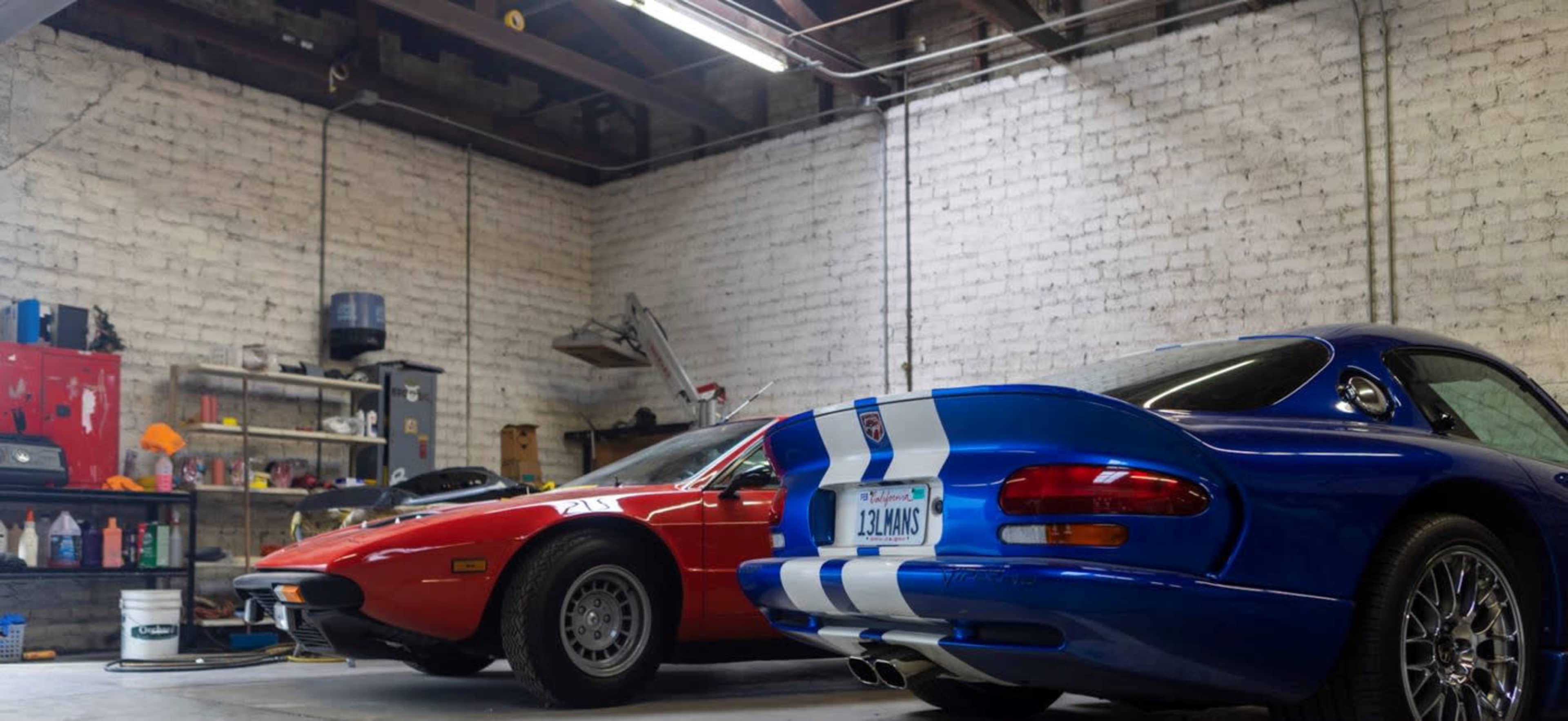 A red sports car and a blue sports car are parked in a garage with exposed brick walls and various tools and supplies organized on shelves.