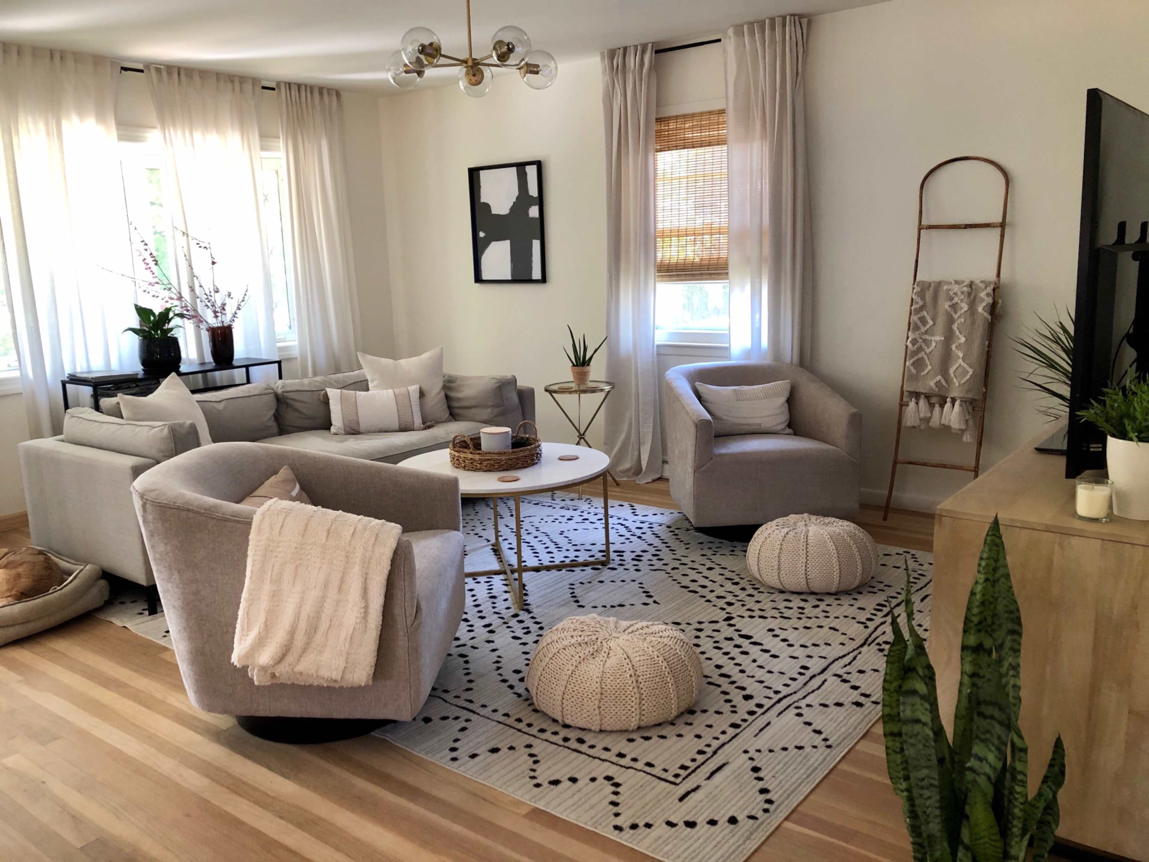 The living room features a mix of light gray furniture, a round coffee table, and decorative plants arranged around a patterned rug.