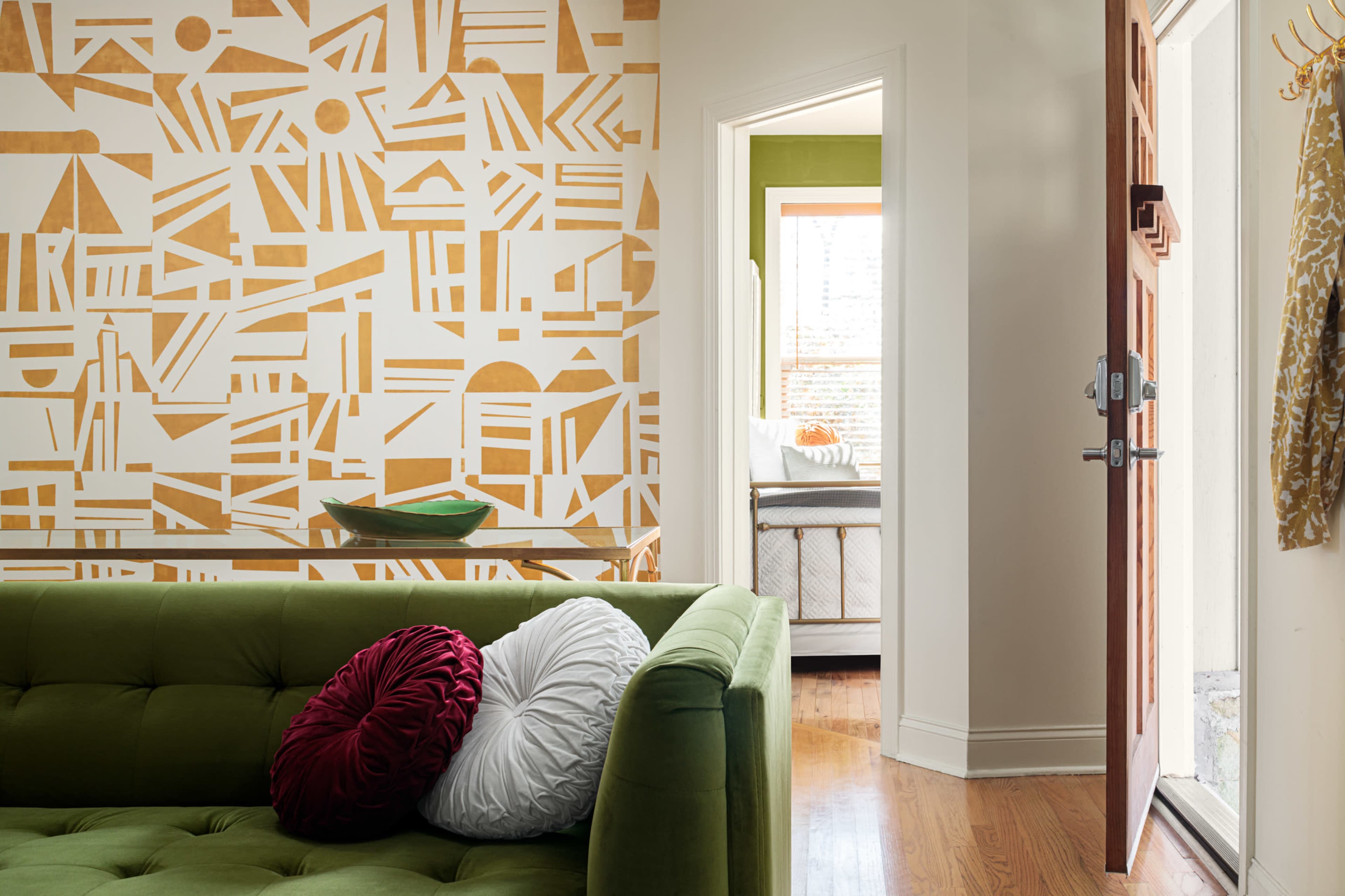 A green couch with decorative pillows in a room featuring a patterned wall and an open doorway leading to another room.