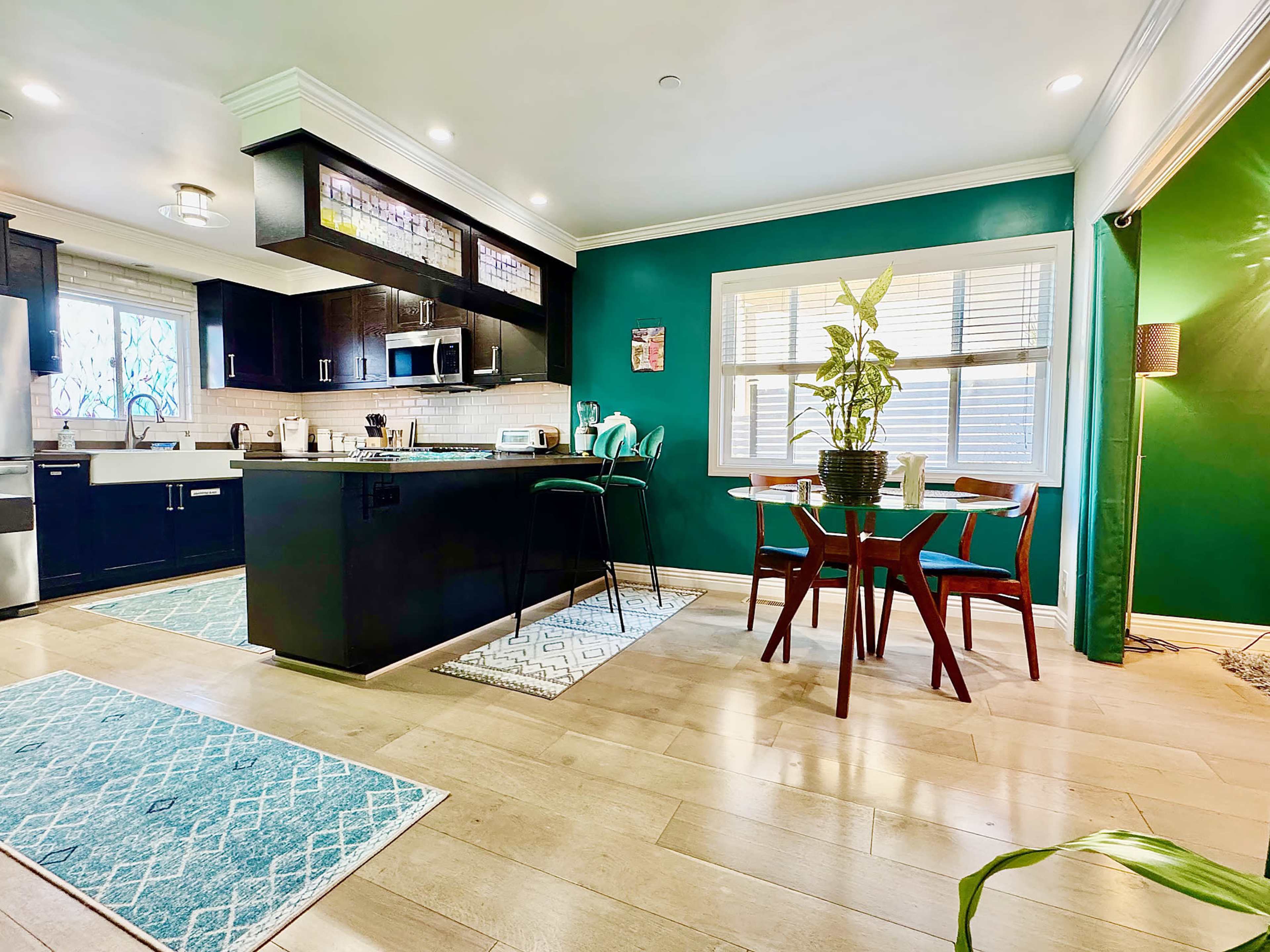 The image shows a modern kitchen with dark cabinets, a green accent wall, and a dining area featuring a small table and chairs.