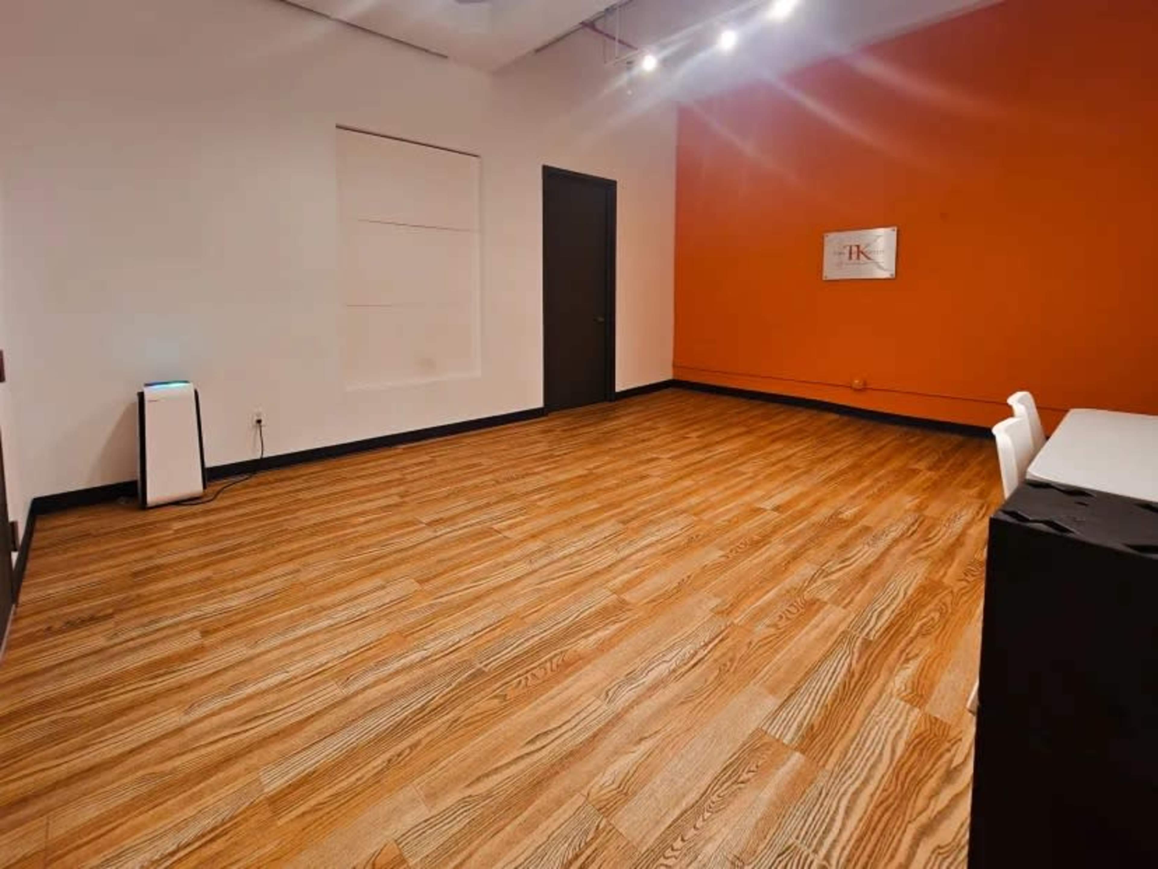 The image shows a spacious room with wooden flooring, an orange accent wall, a door, and minimal furniture, including a white chair and a small appliance.