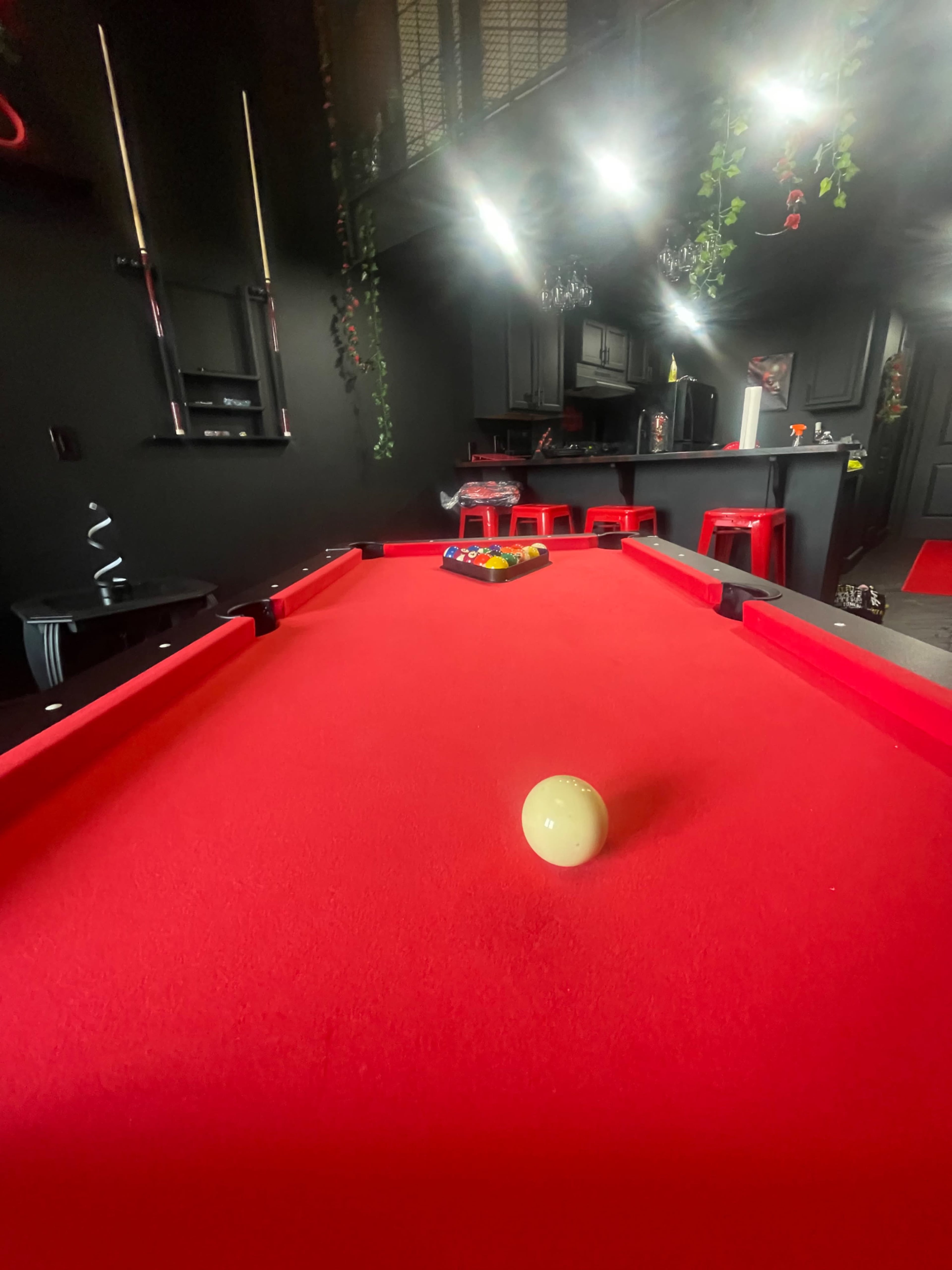 A red felt pool table with a white cue ball is positioned in a dimly lit room featuring black walls and bar-style seating.
