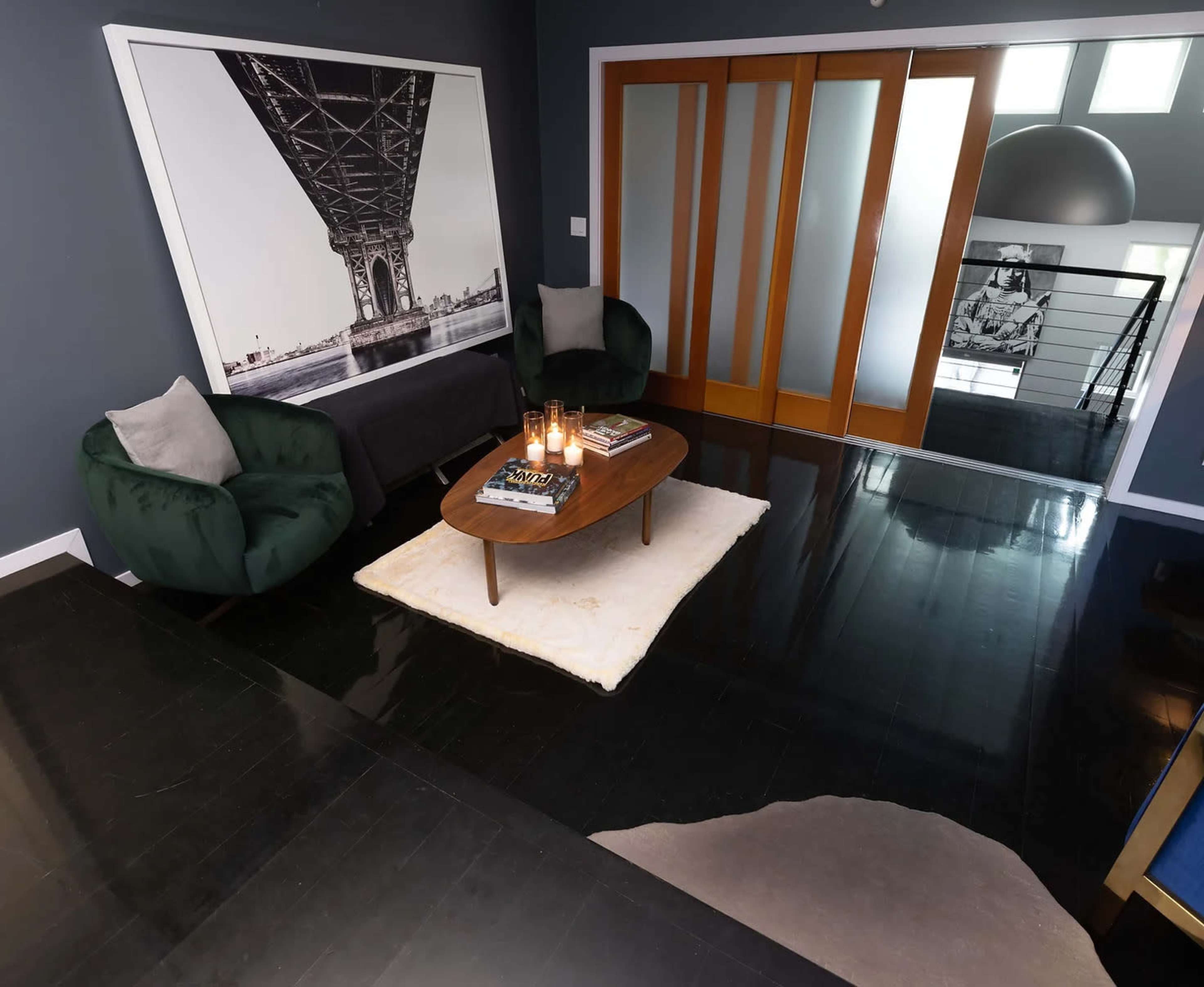 A modern living room features dark hardwood floors, two green armchairs, a coffee table with magazines, and a large wall art piece of a bridge.