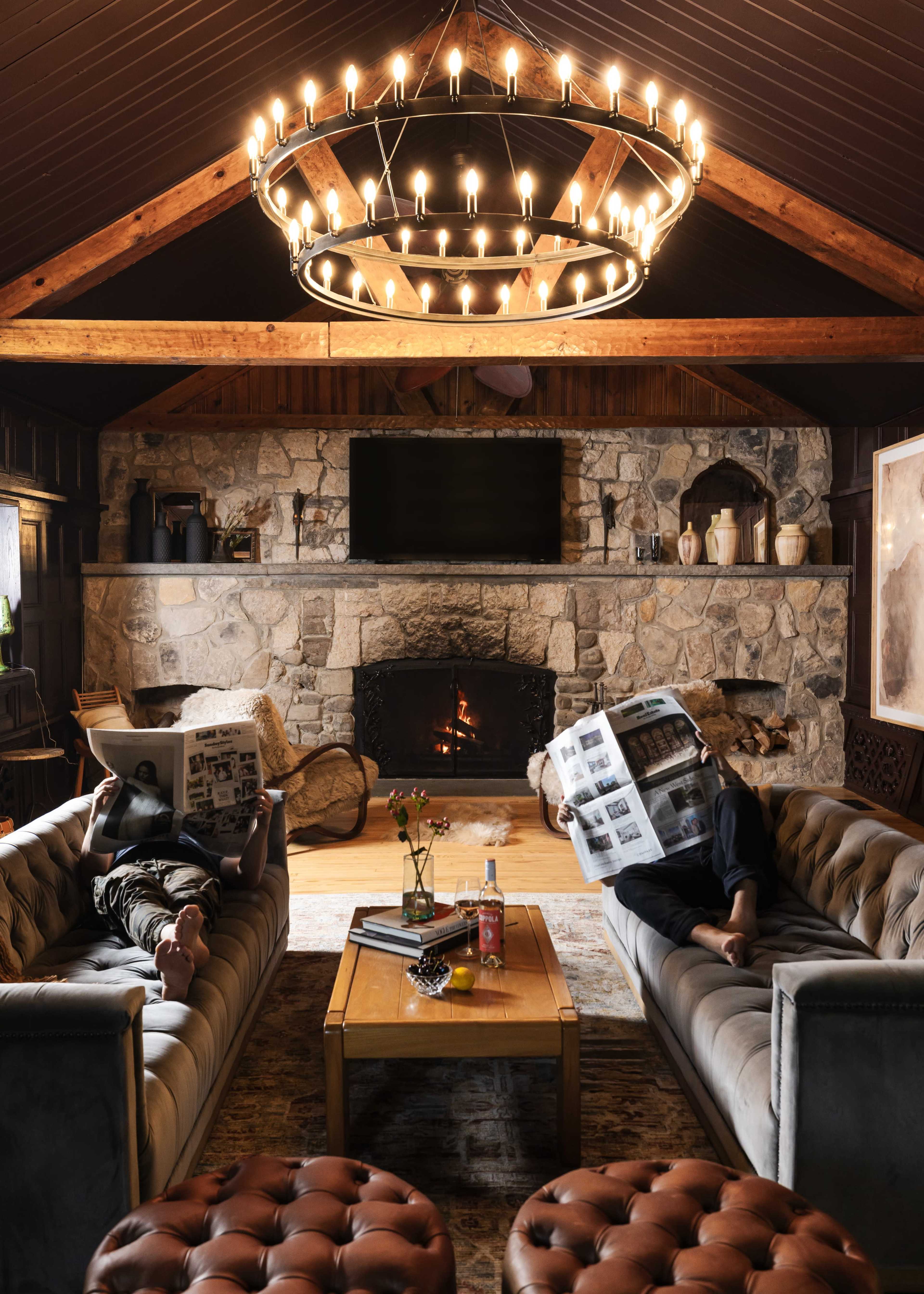 Two people sit on grey sofas in a rustic living room, each reading a newspaper, with a stone fireplace and a large chandelier overhead.