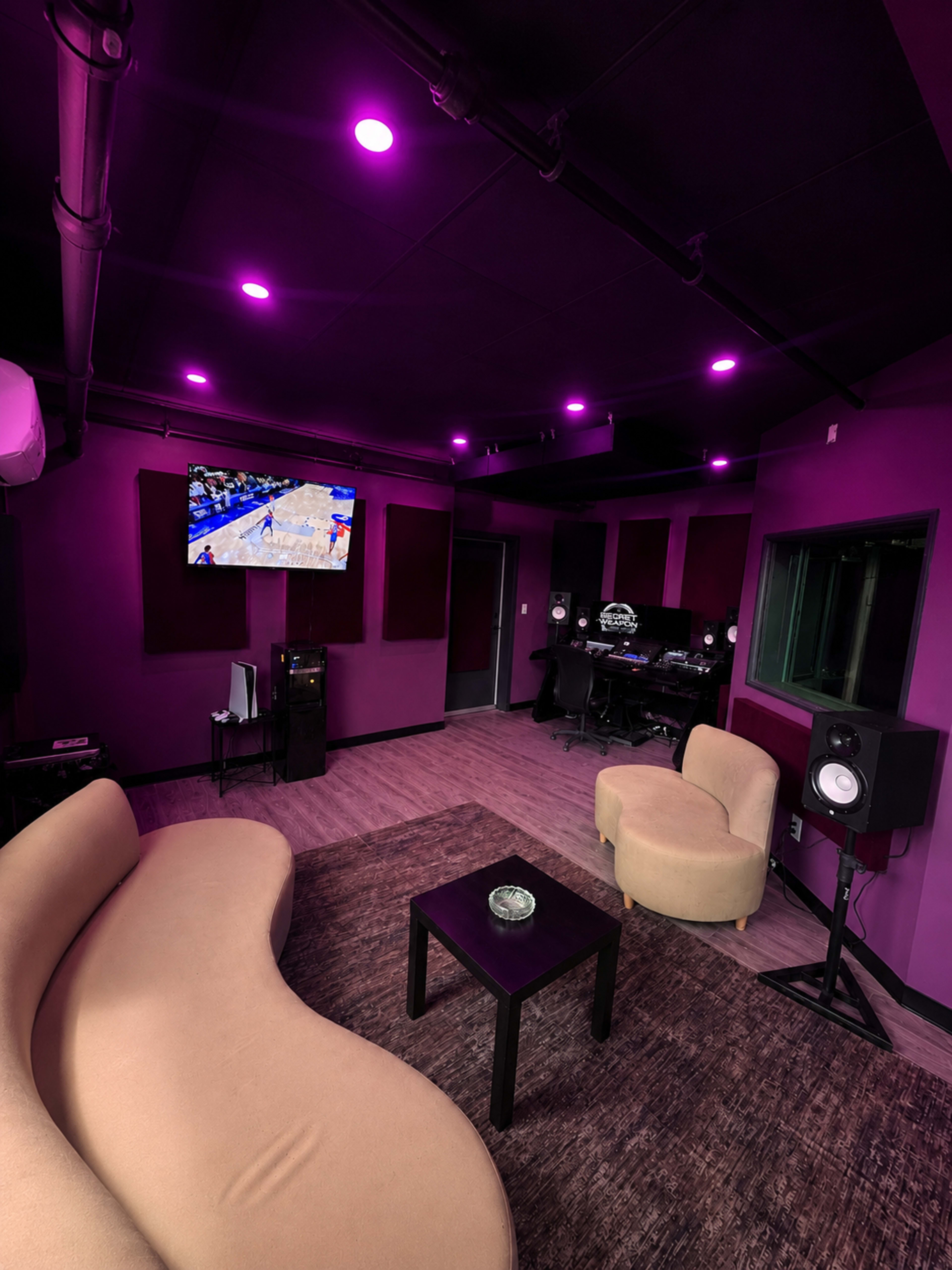 The image shows a modern recording studio with purple walls, two curved sofas, a coffee table, and a screen displaying a sports event.