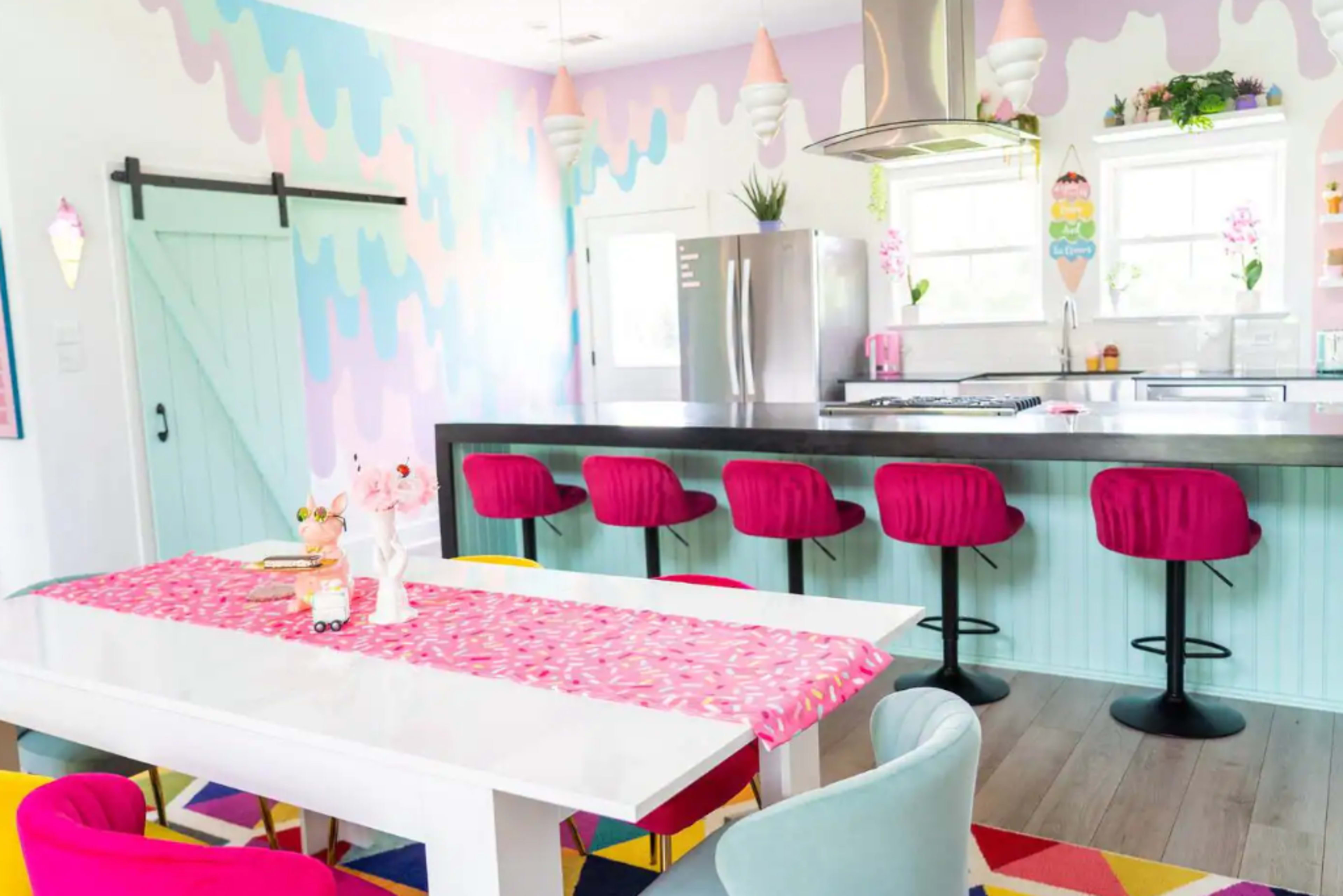 A brightly colored kitchen and dining area with a pastel-themed wall, modern stools at the counter, and a white dining table topped with a vibrant table runner.