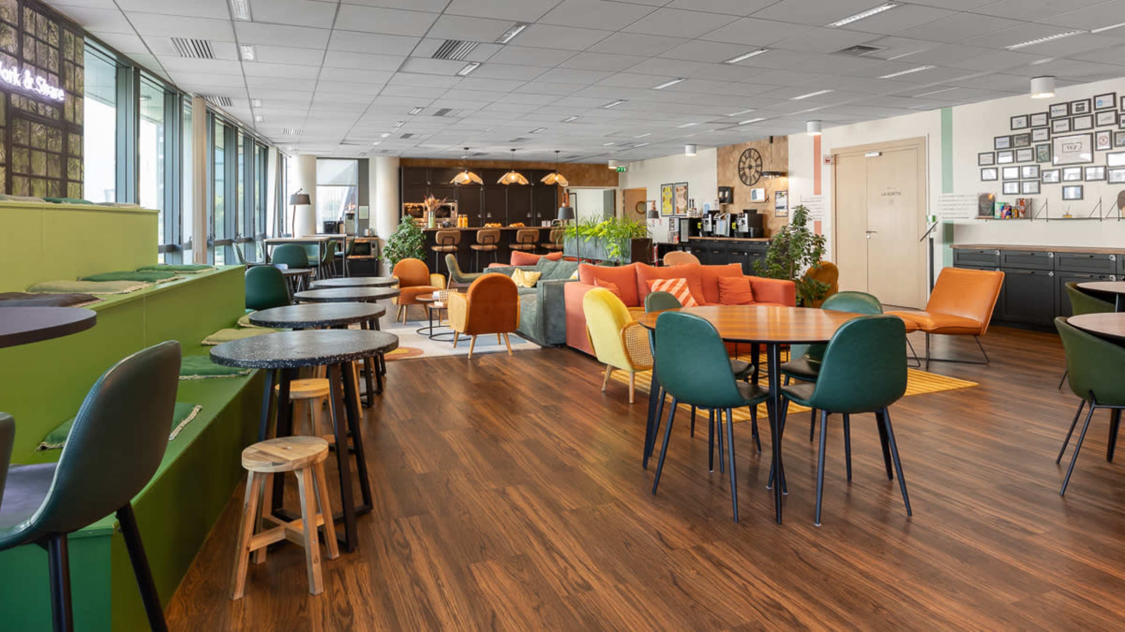 The image shows a spacious modern café with various seating arrangements, including colorful chairs and wooden tables, along with a service area in the background.