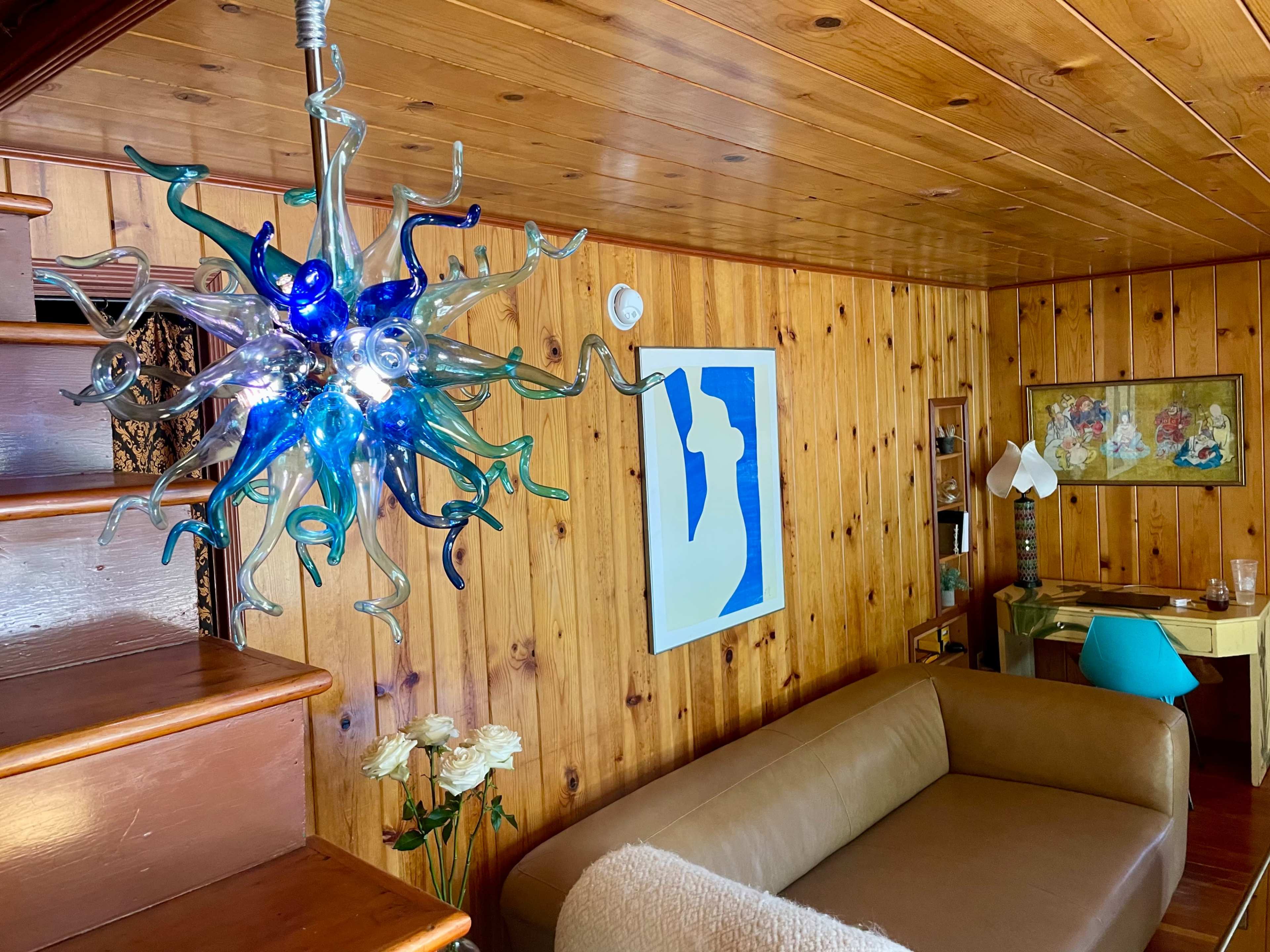 The image shows a living room with a brown couch, a colorful glass chandelier, wooden paneling, and a small desk with a blue chair.