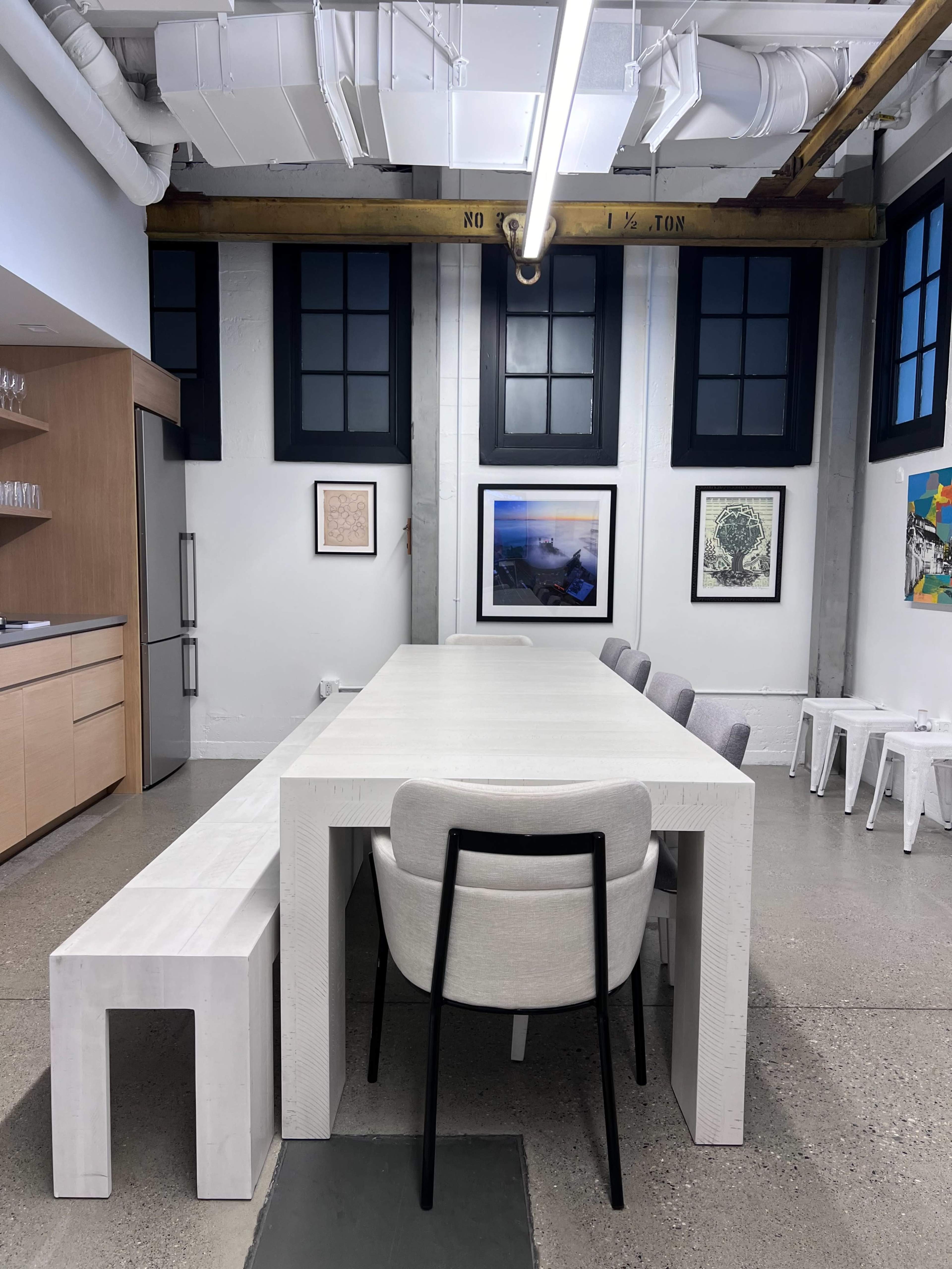 A spacious kitchen area with a long white table, surrounded by gray chairs, and large windows letting in natural light.
