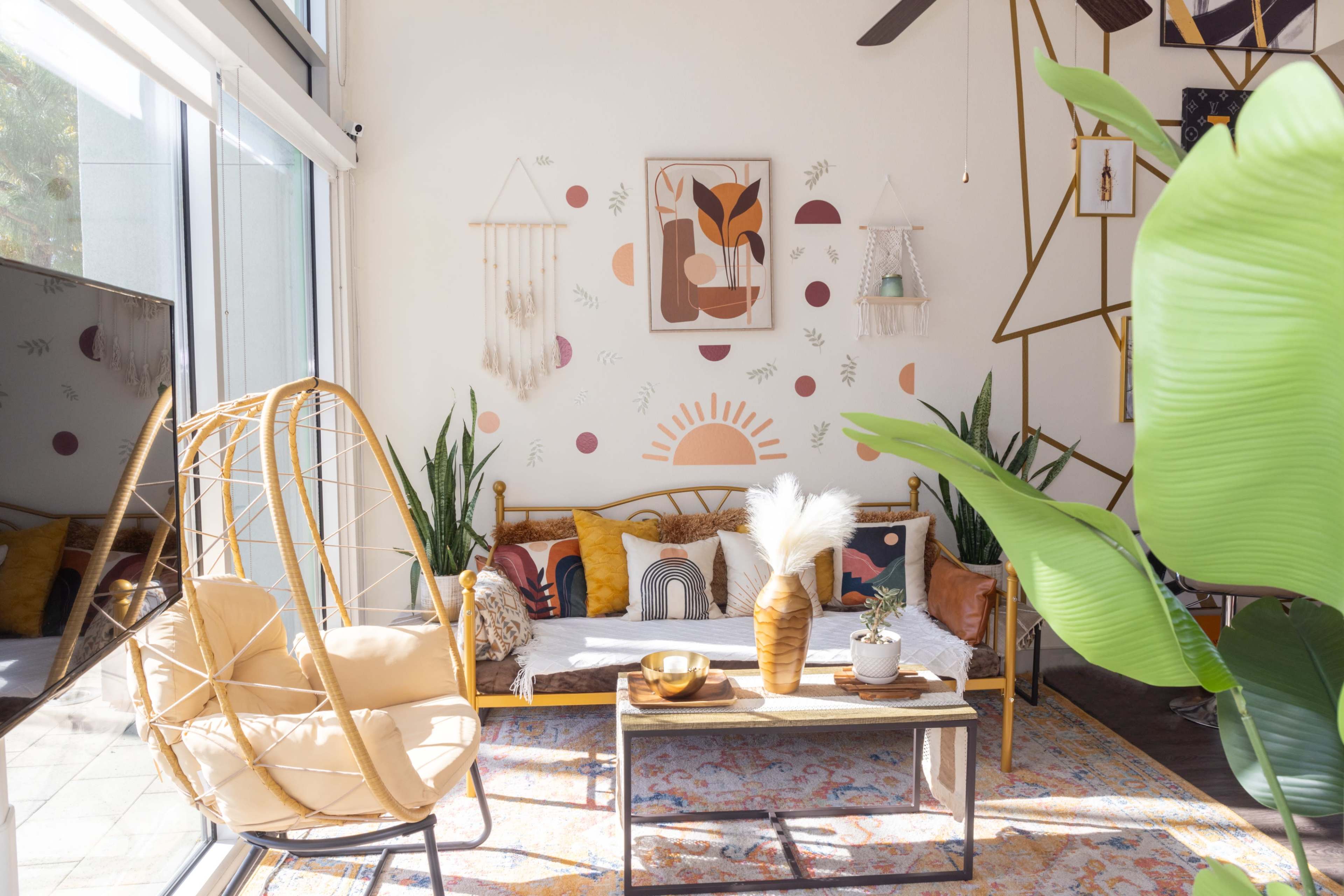 A bright living room with a rattan chair, a sofa adorned with colorful pillows, and decorative wall art featuring geometric patterns and a sun motif.