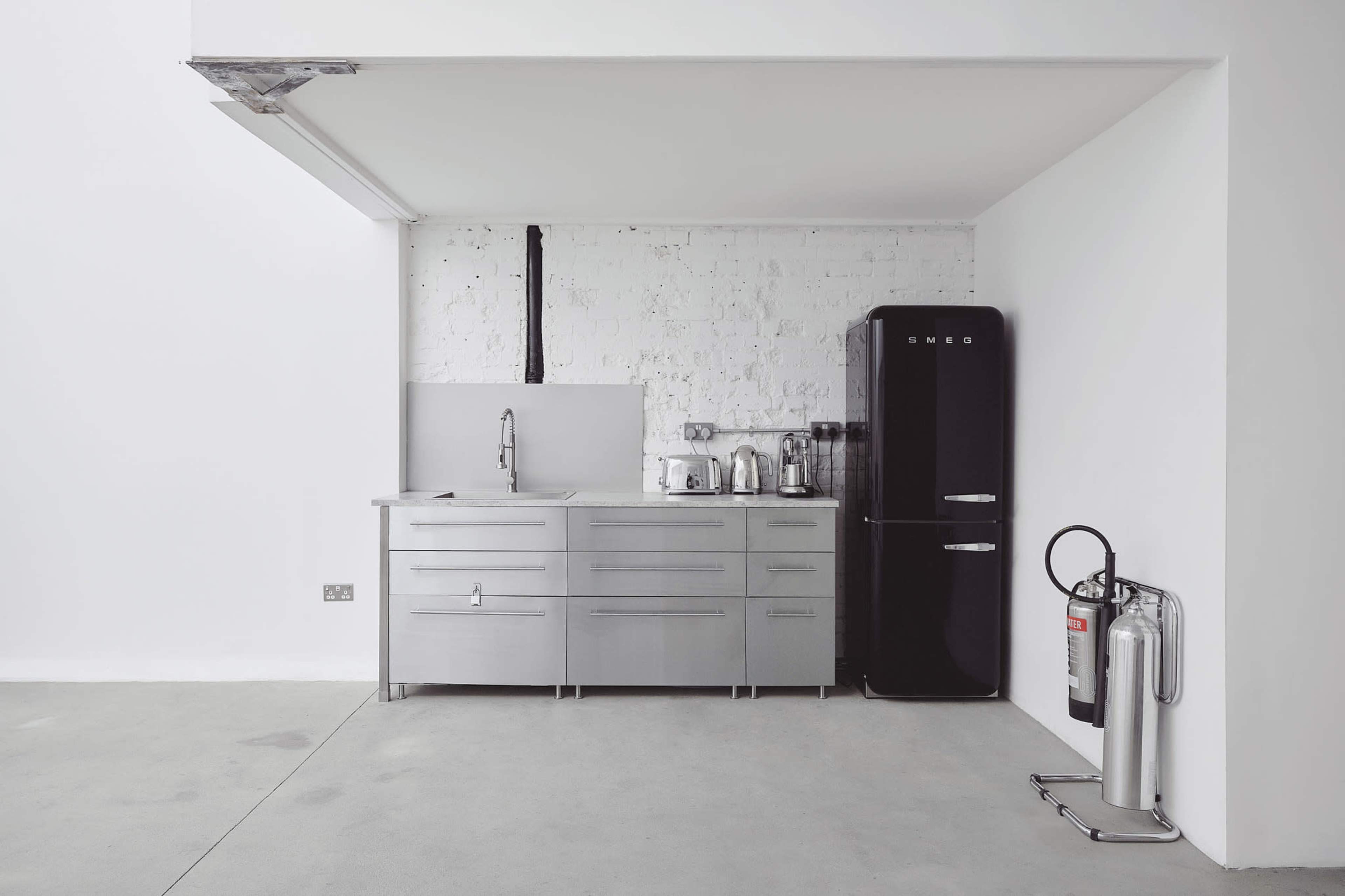 The image shows a minimalist kitchen featuring stainless steel cabinetry, a black refrigerator, and a fire extinguisher mounted on the wall.