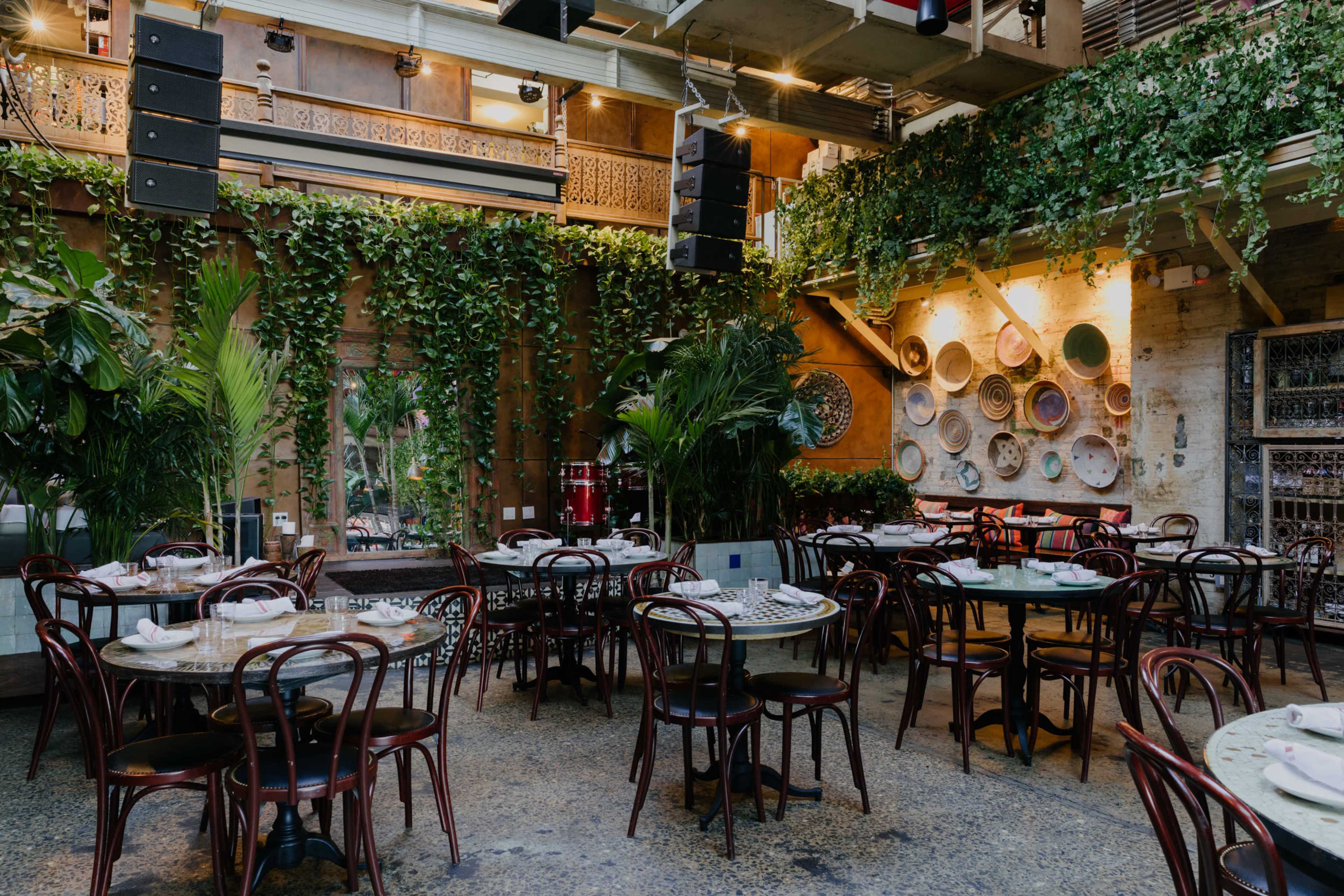 A restaurant interior features round tables with chairs, surrounded by greenery and decorative wall plates, set in a space with industrial decor.