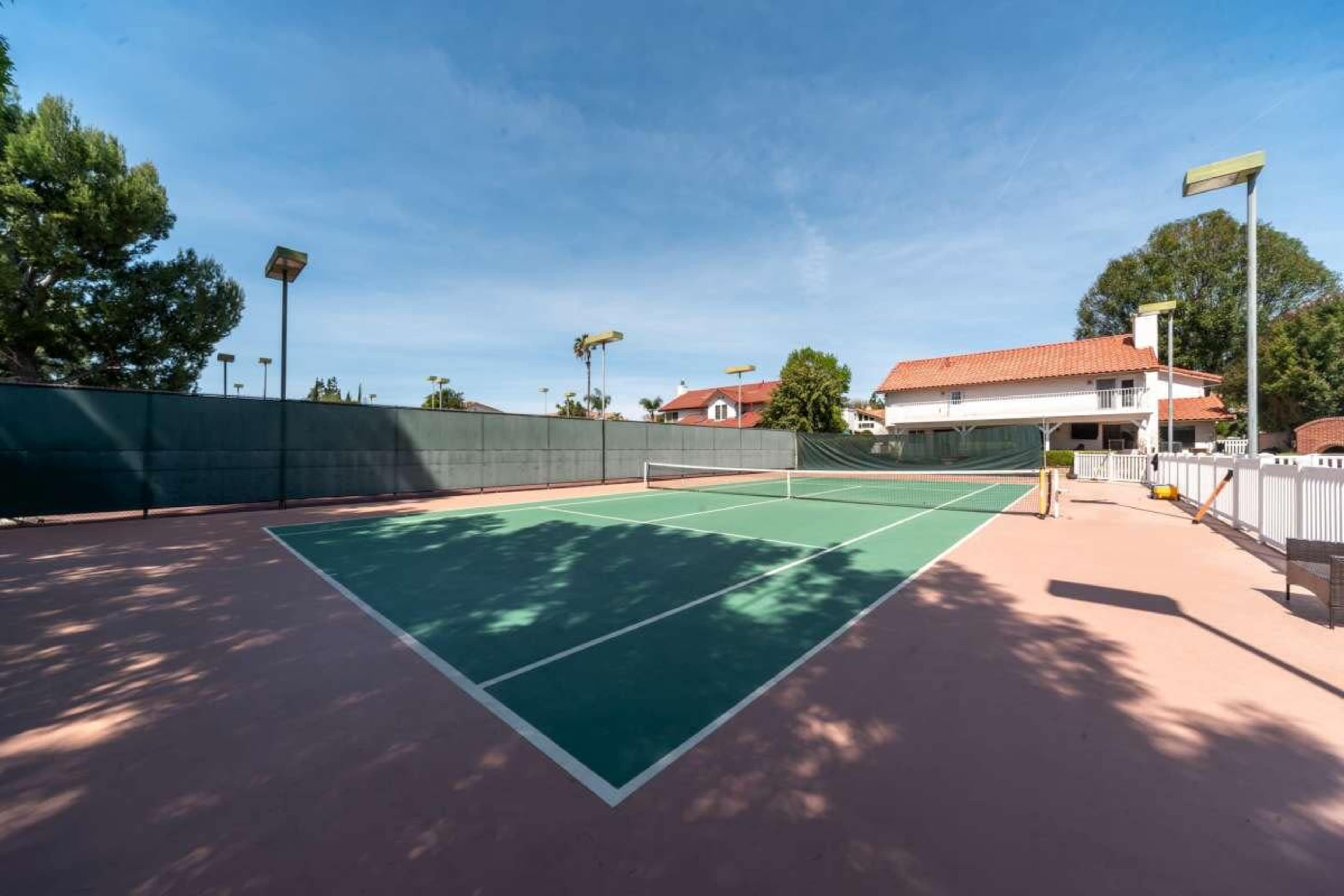 A tennis court with green and pink surfaces is set against a clear blue sky and surrounded by trees and buildings.
