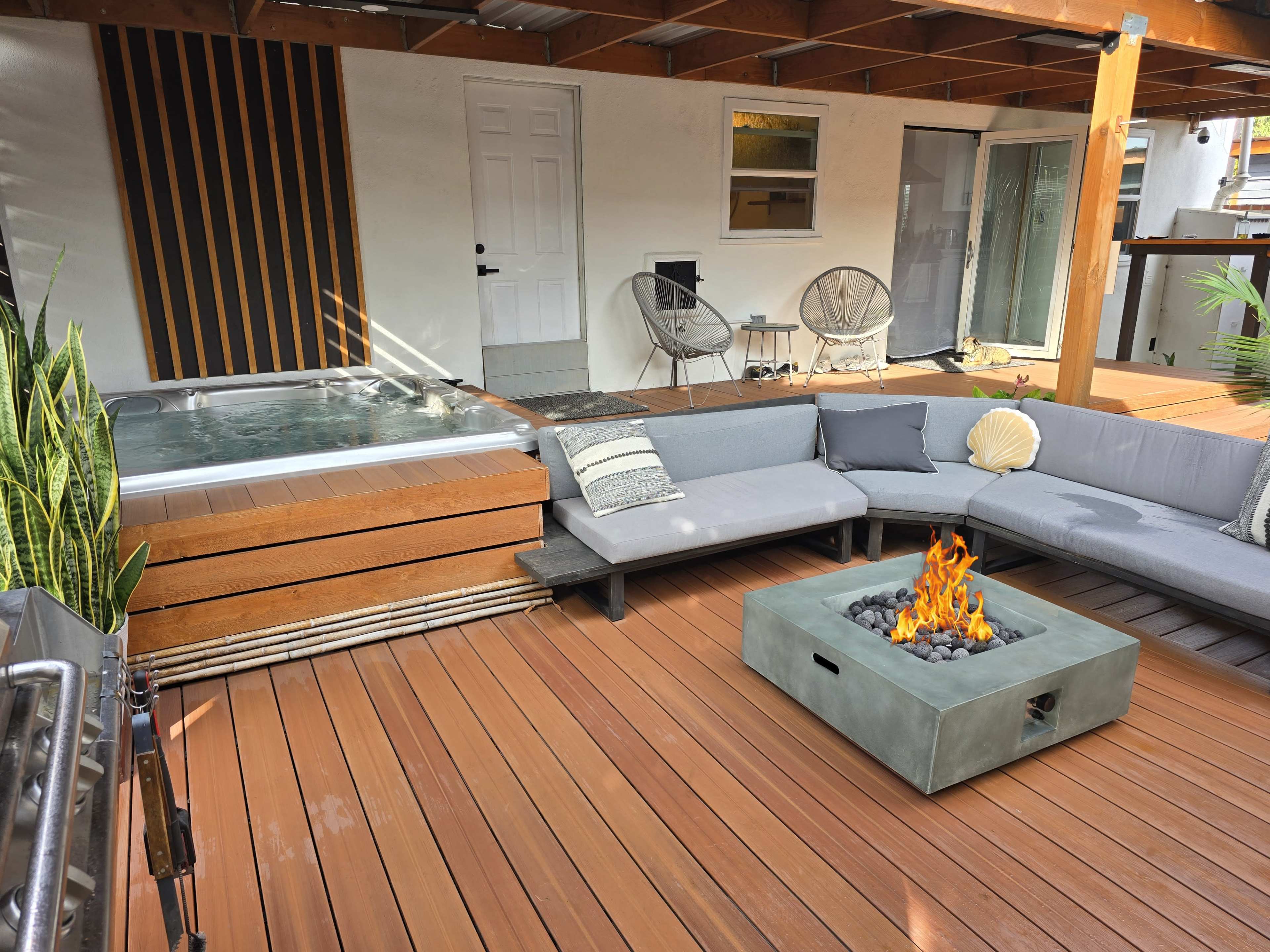 The scene features a modern outdoor lounge area with a sectional sofa, a fire pit, a hot tub, and wooden decking under a covered structure.