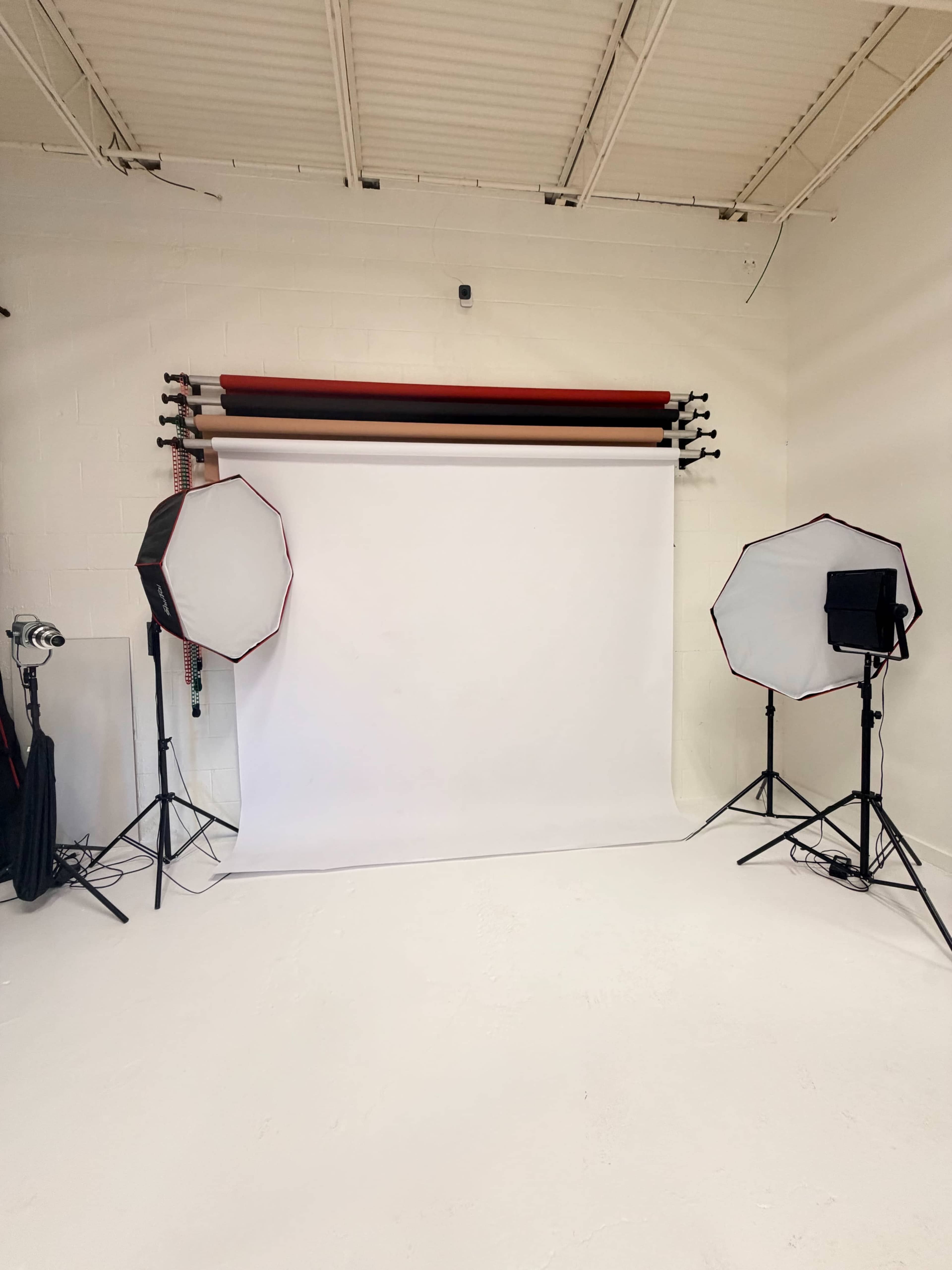 The image shows a photography studio with a white backdrop, two softbox lights on either side, and colored backdrop rolls mounted on the wall.